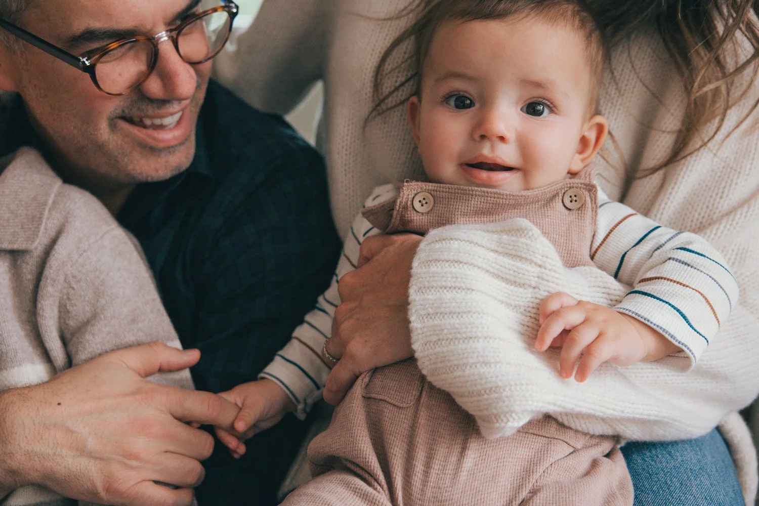 Los Angeles Family photographer. A baby with light hair and big blue eyes, wearing a beige and striped long-sleeve shirt and overalls, being held by a woman while a man nearby reaches out and touches the baby's hand. The scene is warm and joyful.