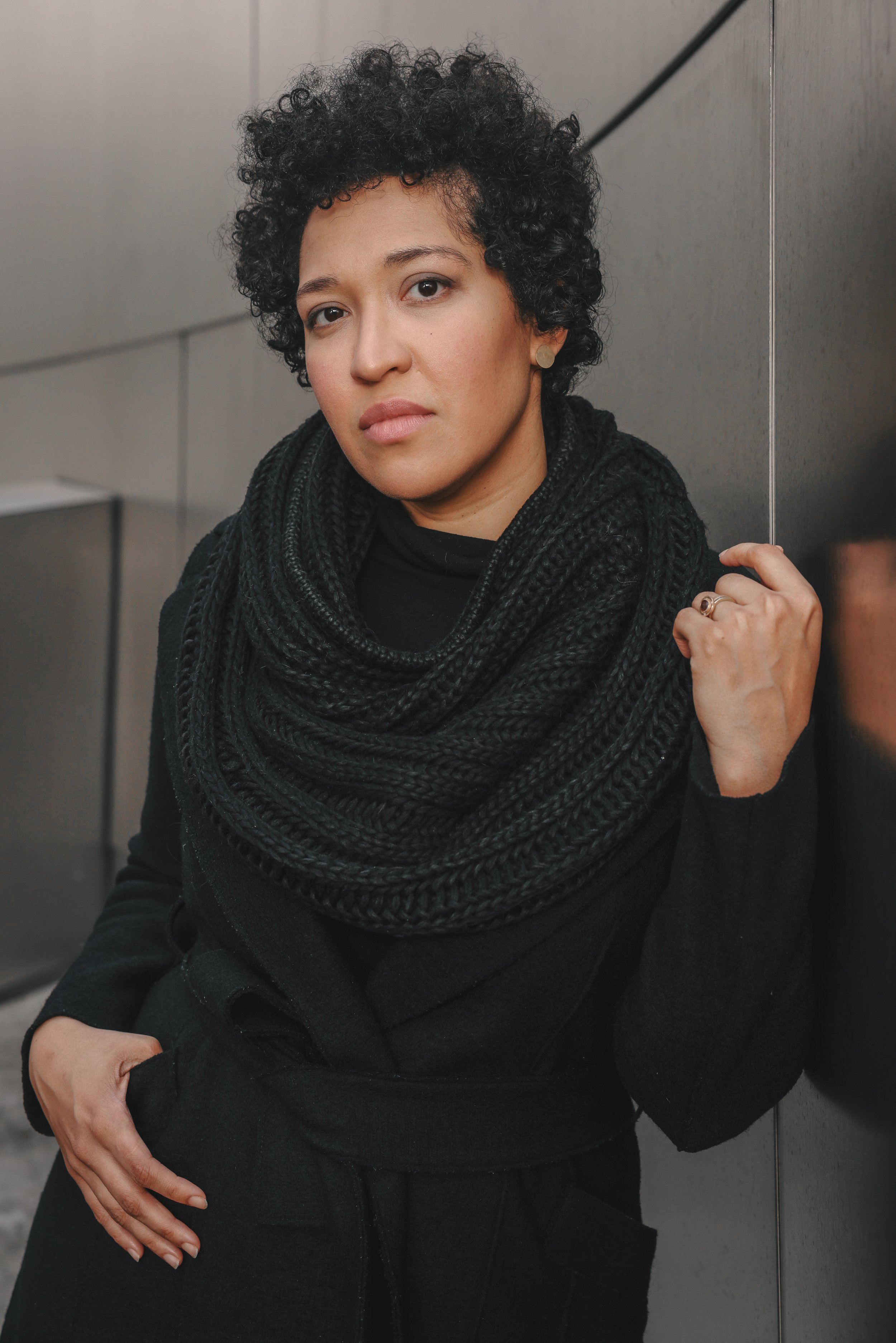 Close-up portrait of a person with short curly black hair wearing a black sweater and chunky scarf, posing indoors.