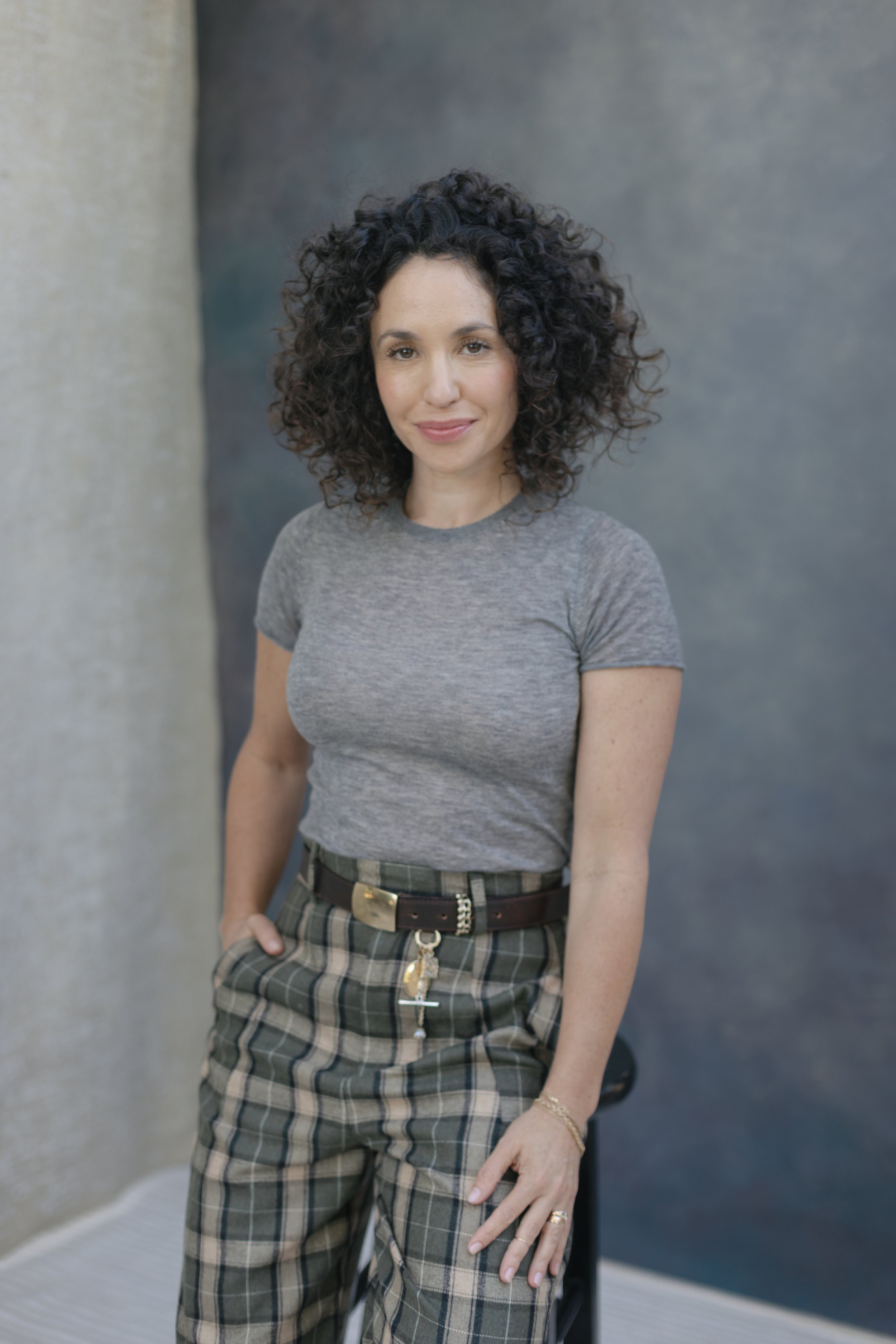 A woman with dark, curly hair smiling, wearing a gray t-shirt and plaid pants, standing outdoors with a gray and beige textured wall in the background.