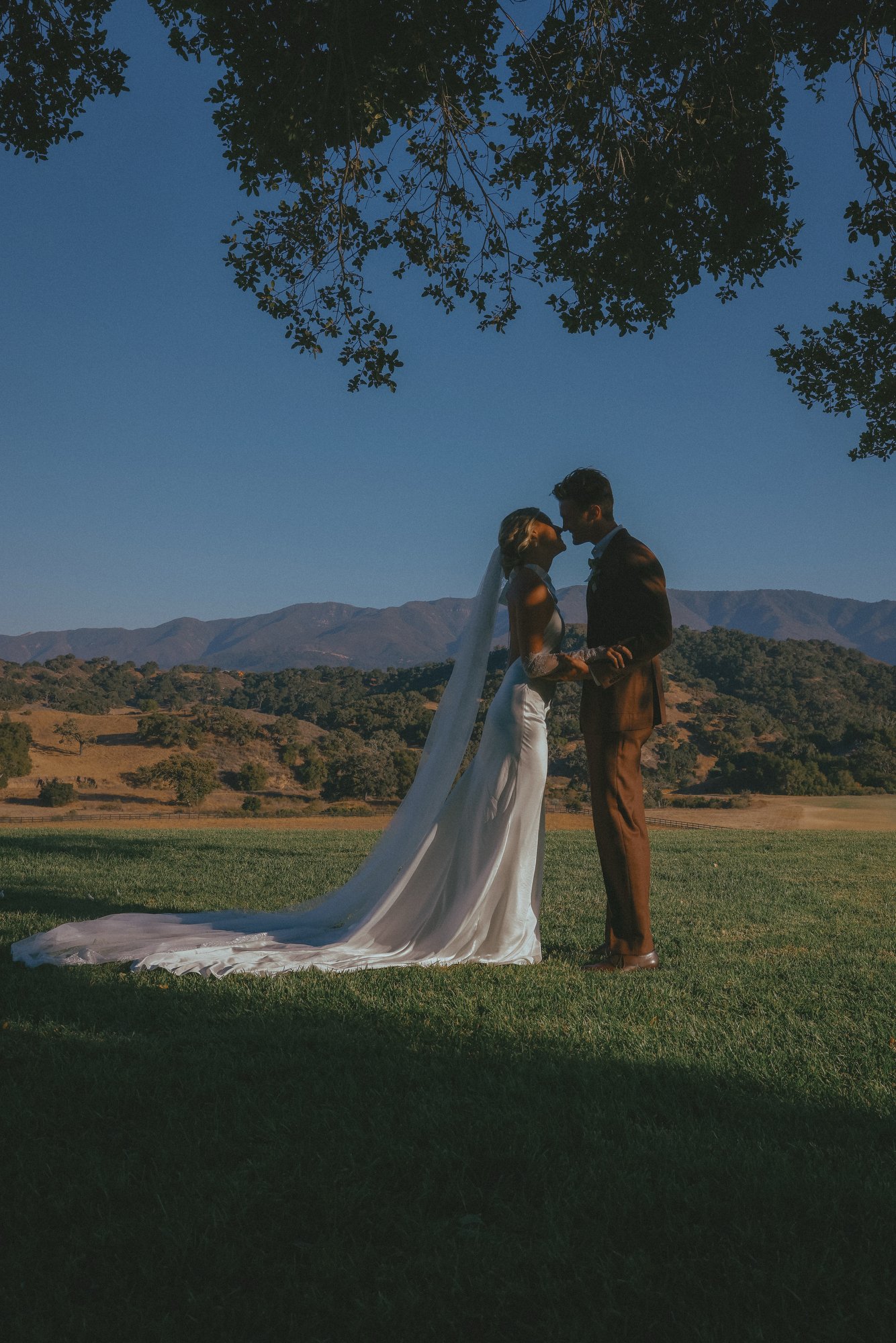 A bride and groom holding hands and about to kiss outdoors in a grassy field with mountains in the background.