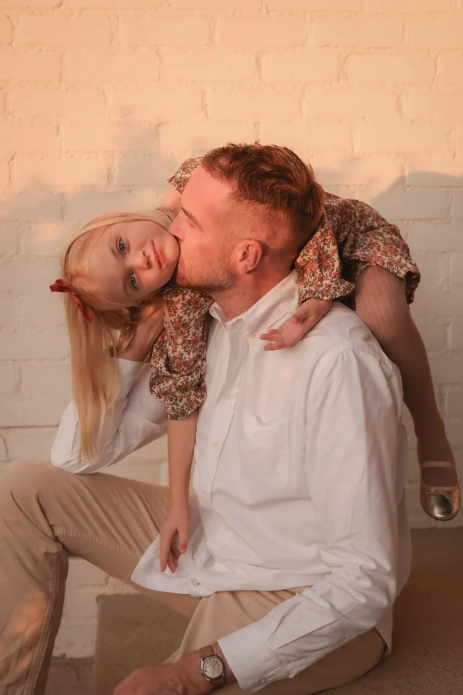 A man sitting on the floor with a young girl on his shoulders, giving her a kiss on the cheek. The girl has long blonde hair with a red bow and is wearing a floral dress. They are in front of a white brick wall.