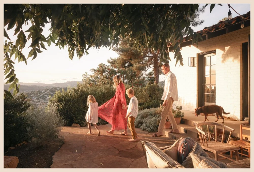 Family walking outside near a white house with a dog on the porch during sunset, with greenery and hills in the background.