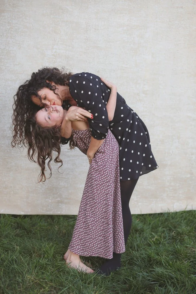 Two women sharing a kiss, standing on grass in front of a plain wall. One woman wears a black dress with white polka dots and black tights, while the other wears a pink patterned dress and is barefoot.