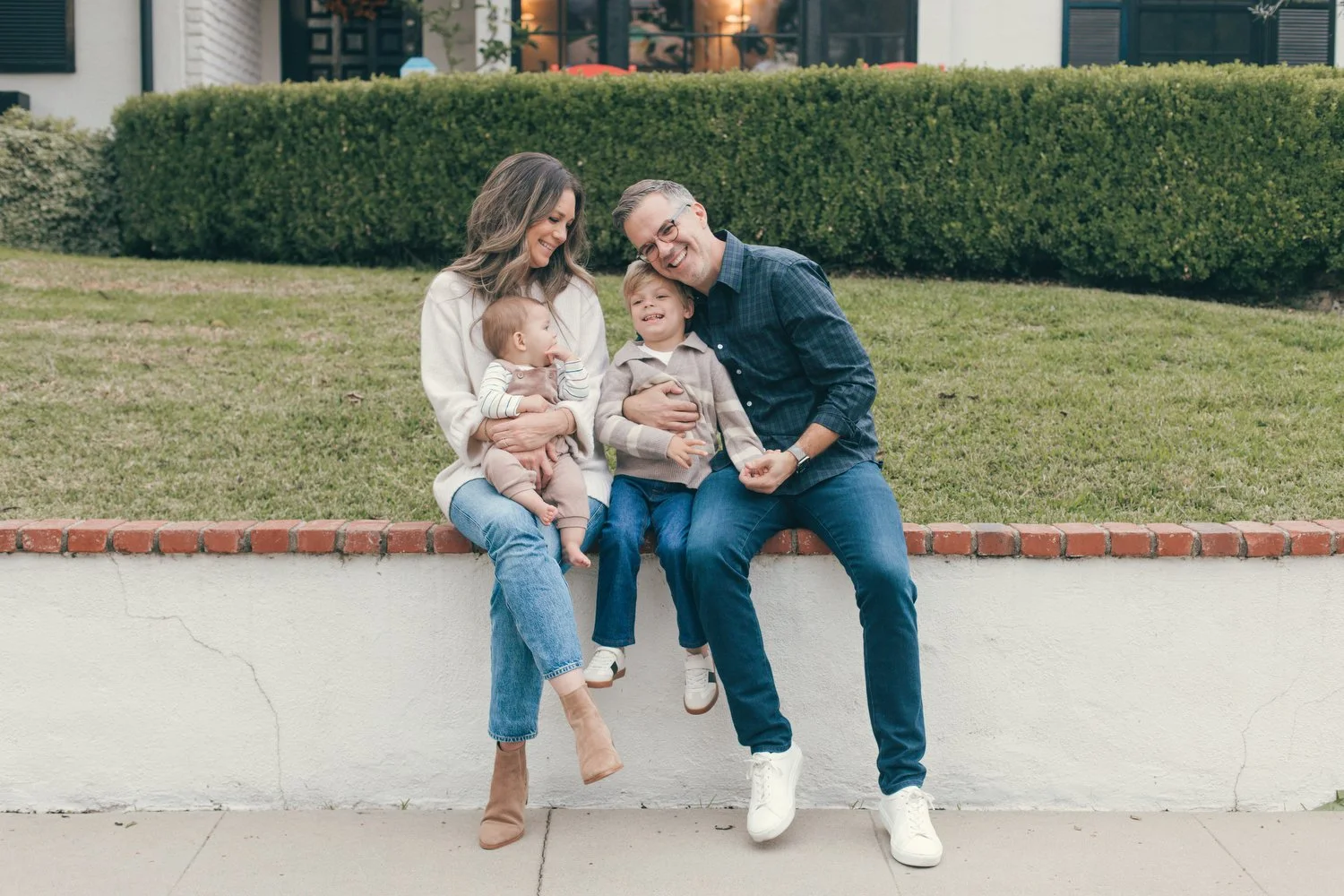 Los Angeles Family photographer. Santa Barbara family photographer. A family of four sitting on a low wall outdoors, smiling and enjoying each other's company. The family includes a woman, a man, a young boy, and a baby.