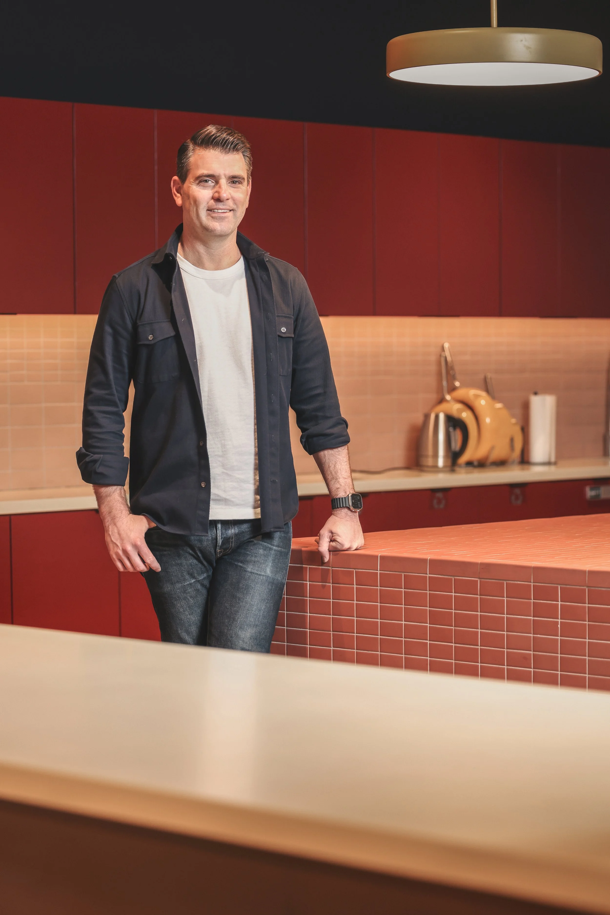Smiling man in a black overshirt standing in a modern kitchen with red tile walls and pendant lighting