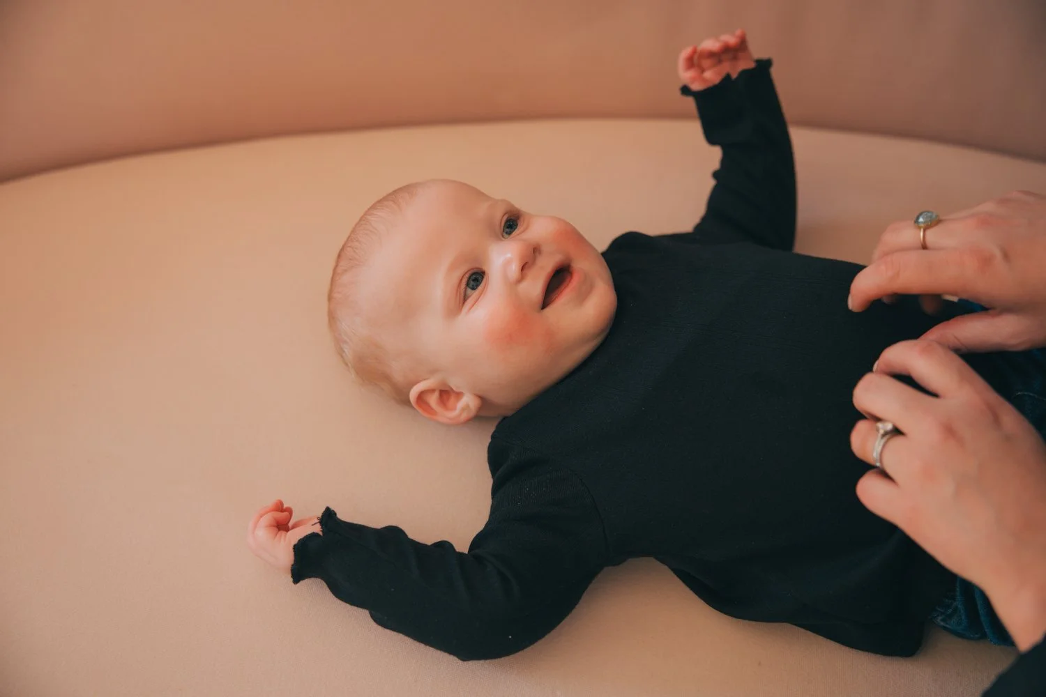 Los Angeles Family photographer. Santa Barbara family photographer.  Newborn. A smiling baby lying on a beige cushion, wearing a black long sleeve shirt, with hands raised. An adult's hands are touching the baby's stomach, and the baby is looking up.