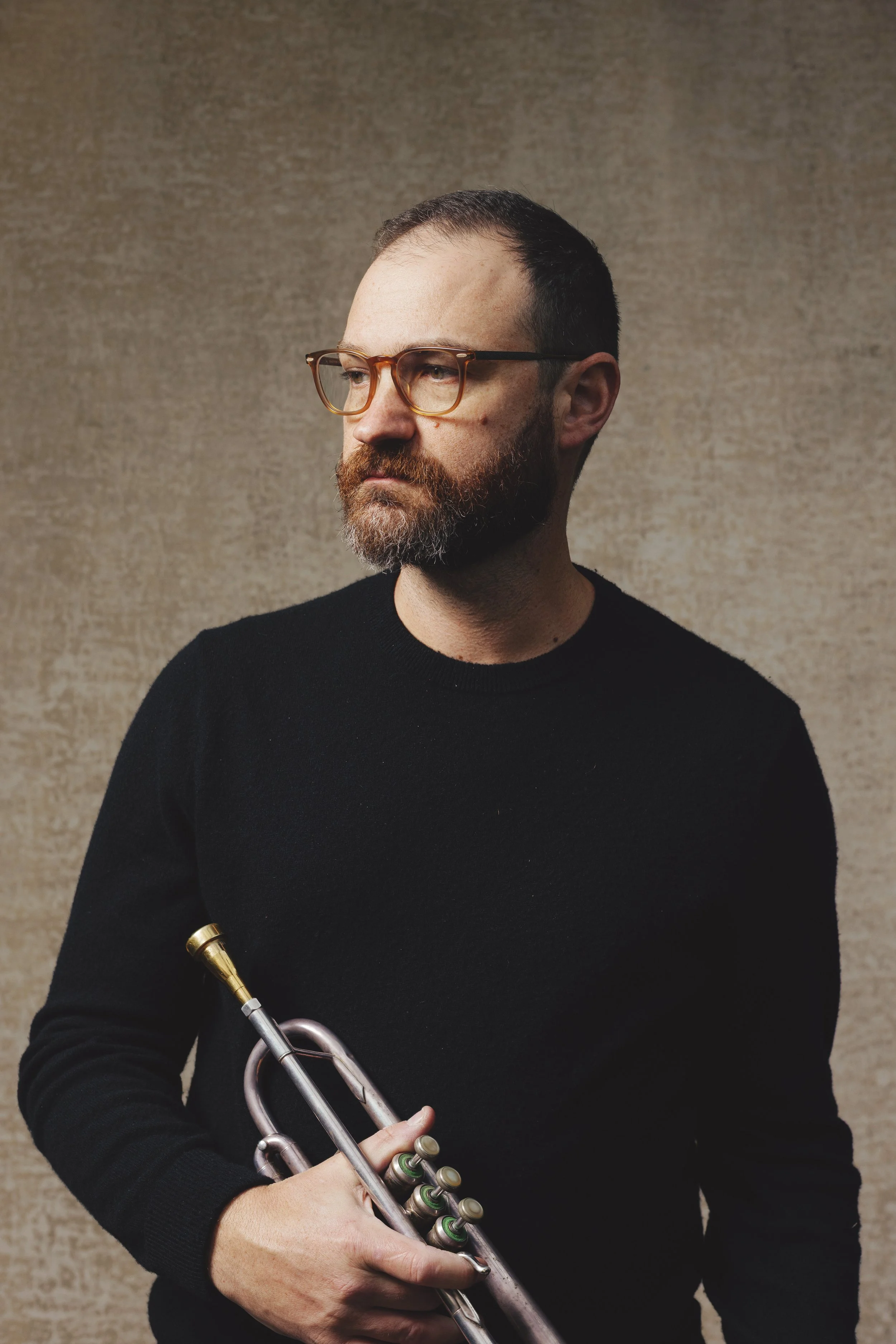 A man with glasses and a beard holding a trumpet against a beige textured background.