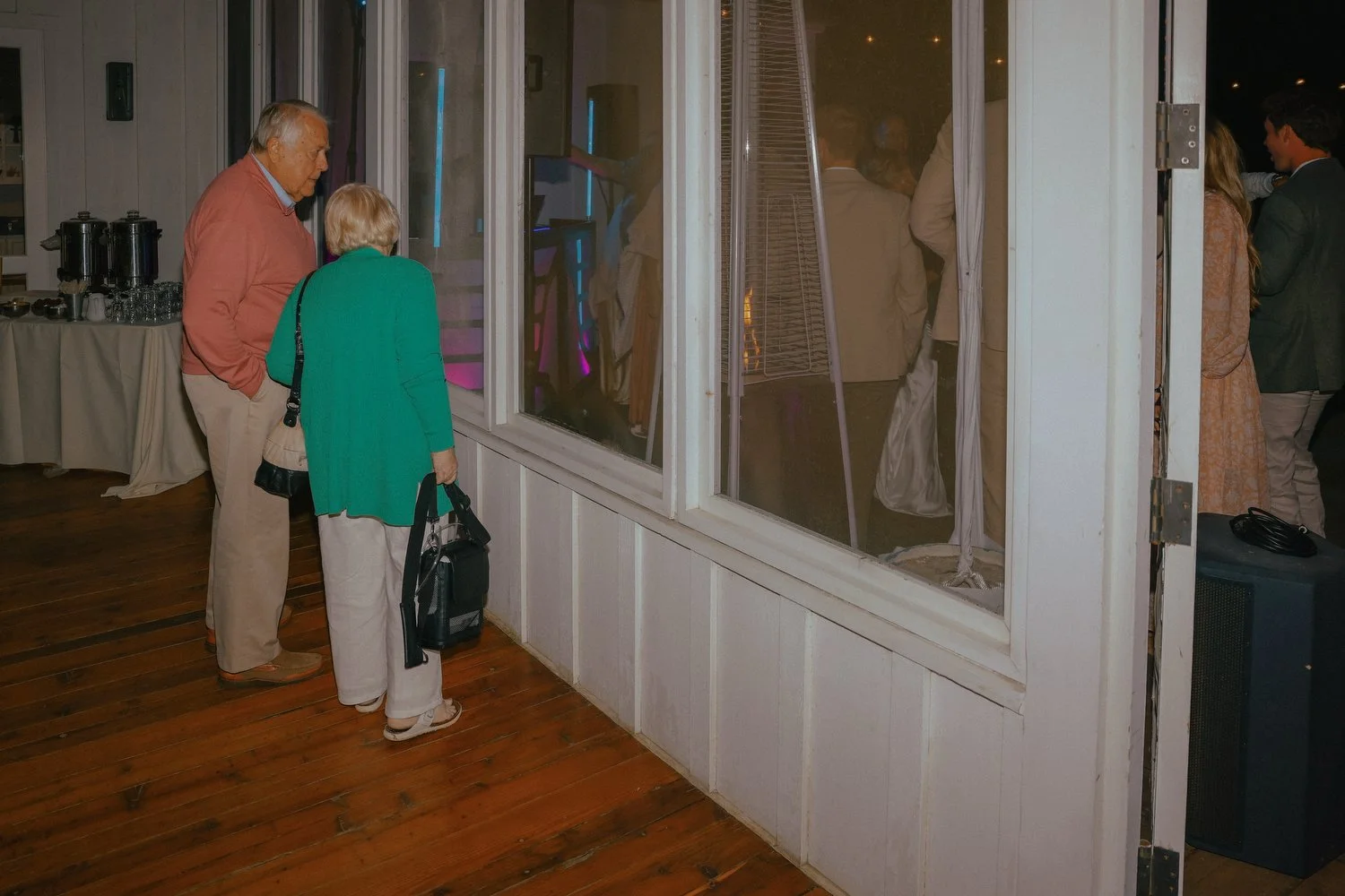An elderly man and woman looking through a window at a wedding inside a room with people standing and talking. Santa Barbara wedding photographer.