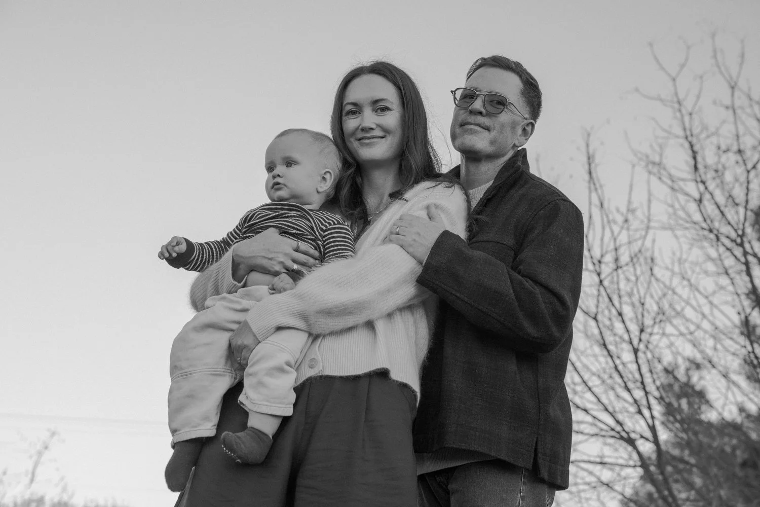 Los Angeles Family photographer. Santa Barbara family photographer.  A family of three standing outdoors in black and white. The mother is holding a young child, and the father is standing beside them.