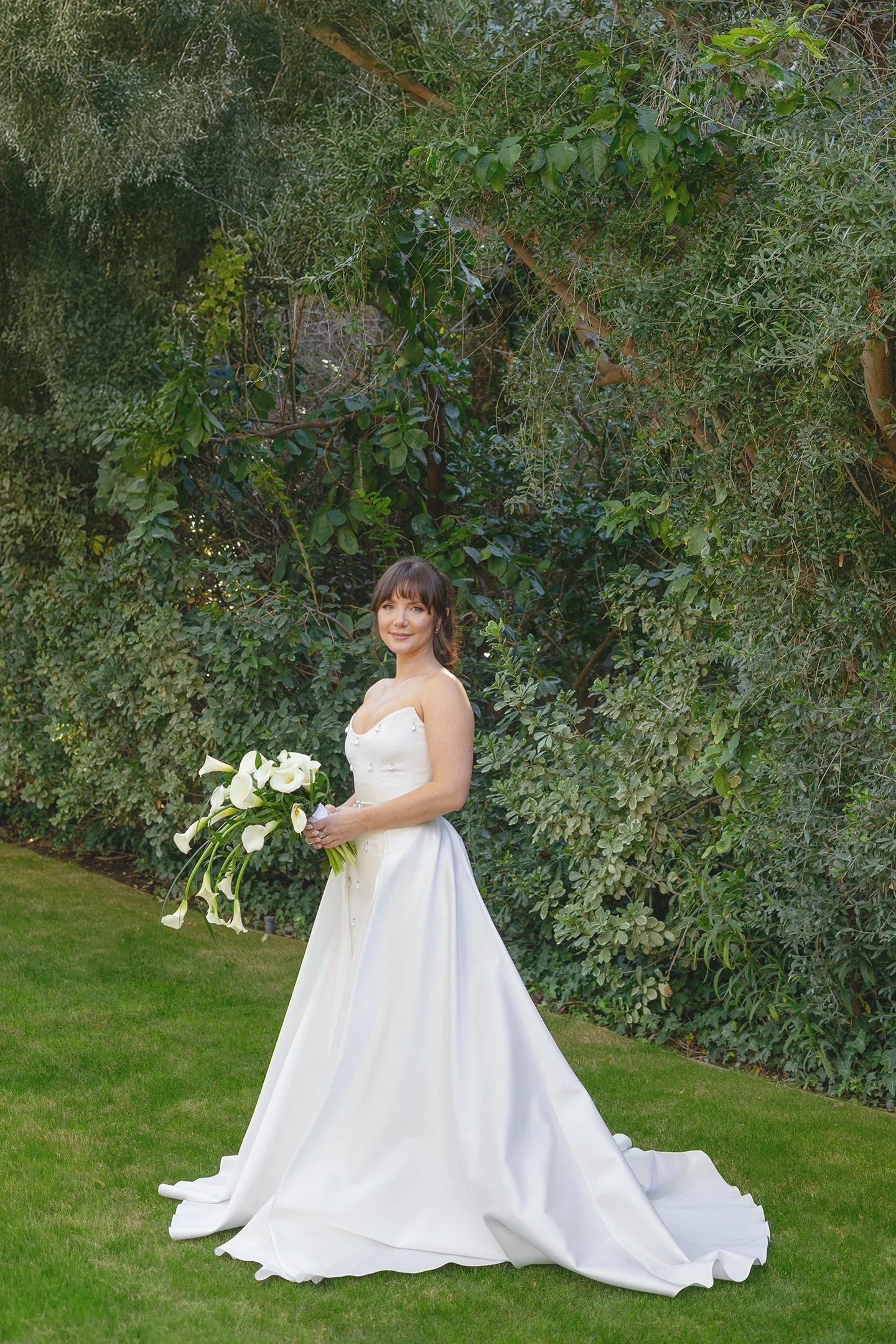A woman in a white wedding dress holding a bouquet of calla lilies, standing on a green lawn with dense trees and foliage in the background.