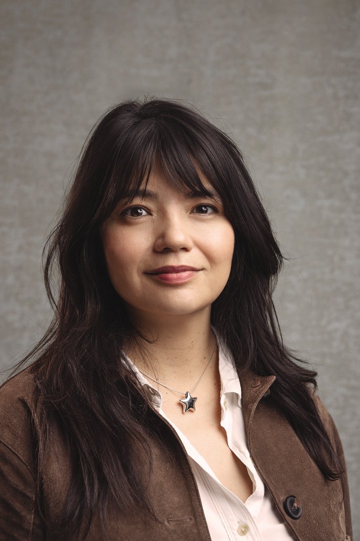 A woman with long dark hair, wearing a brown jacket and a white shirt, standing against a neutral gray background.