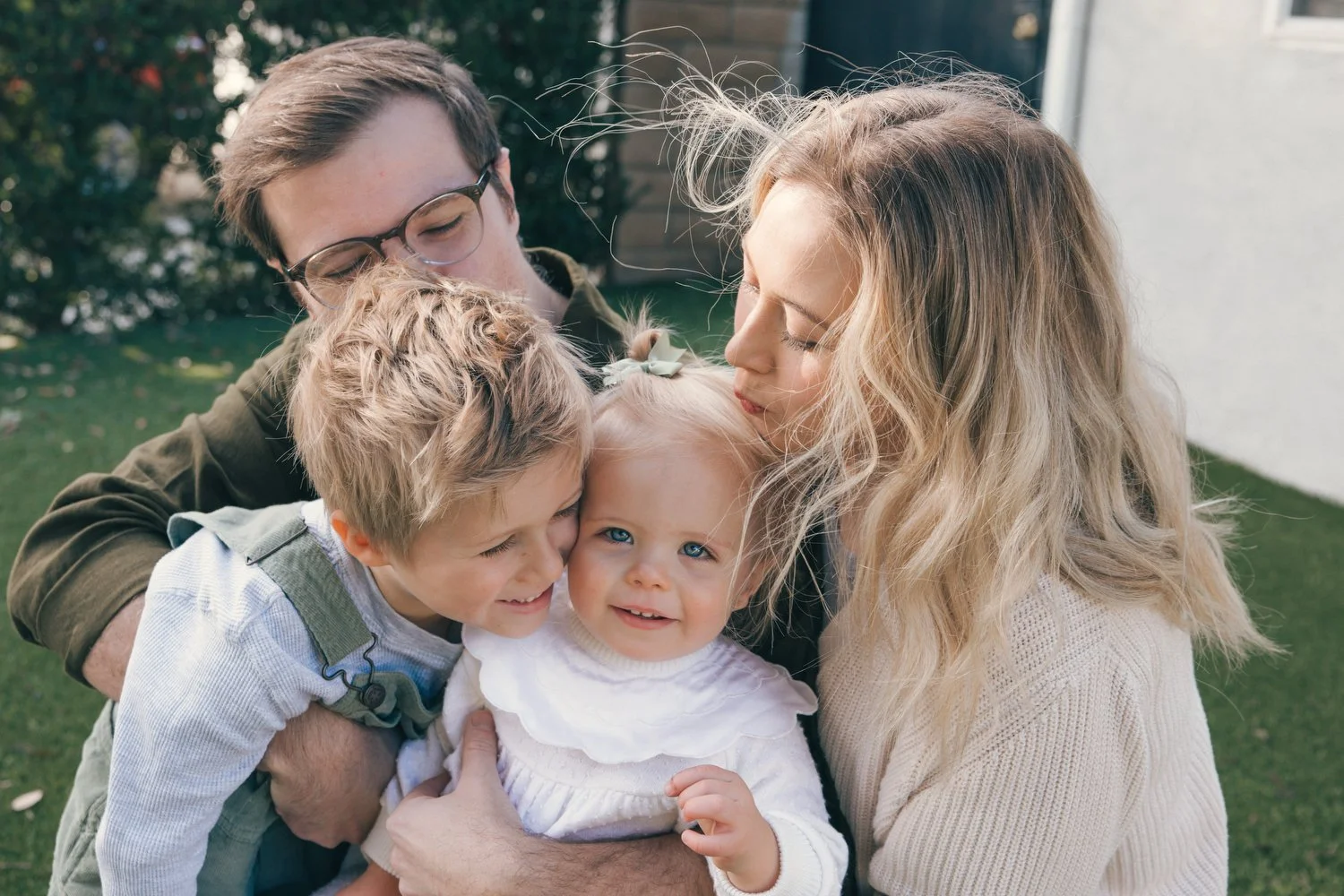 Los Angeles Family photographer. Santa Barbara family photographer.  A family of four, including a man, woman, and two young children, hugging outdoors on a grassy yard with plants and a house in the background.