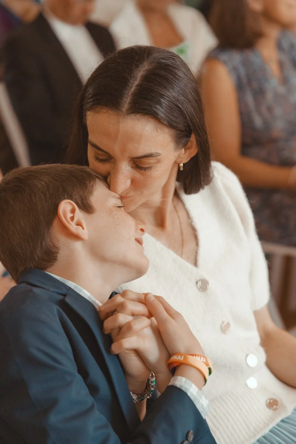 A woman kisses a young boy on the forehead in a crowded setting, holding his hands.
