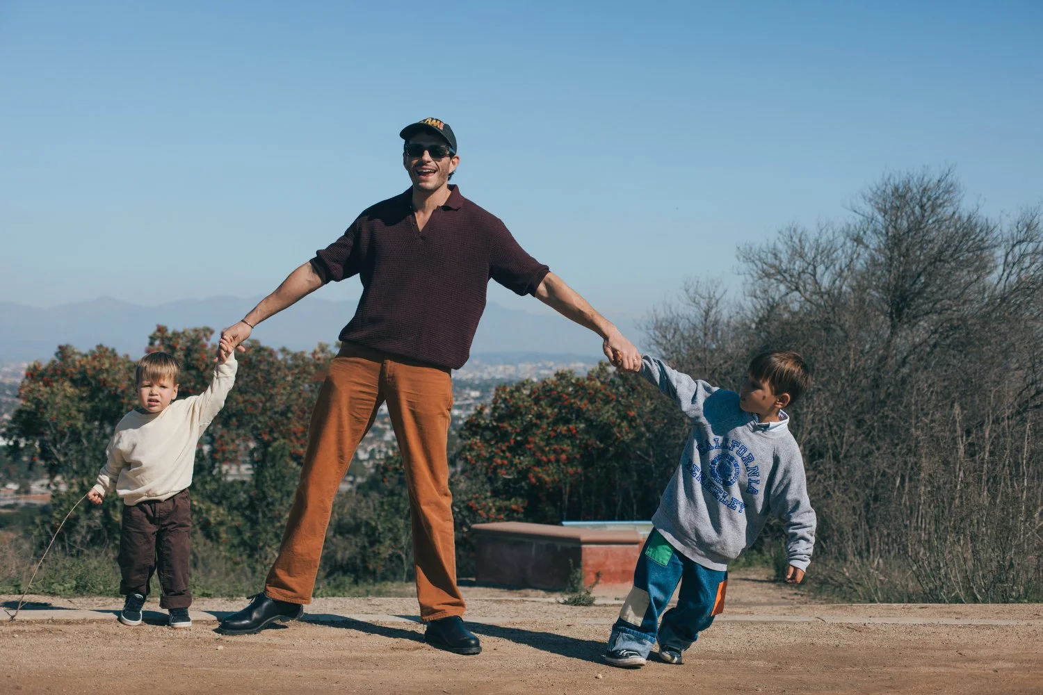 A man holding hands with two young boys outdoors on a dirt path with trees and distant mountains in the background, under a clear blue sky. Los Angeles Family photographer. Santa Barbara family photographer. 