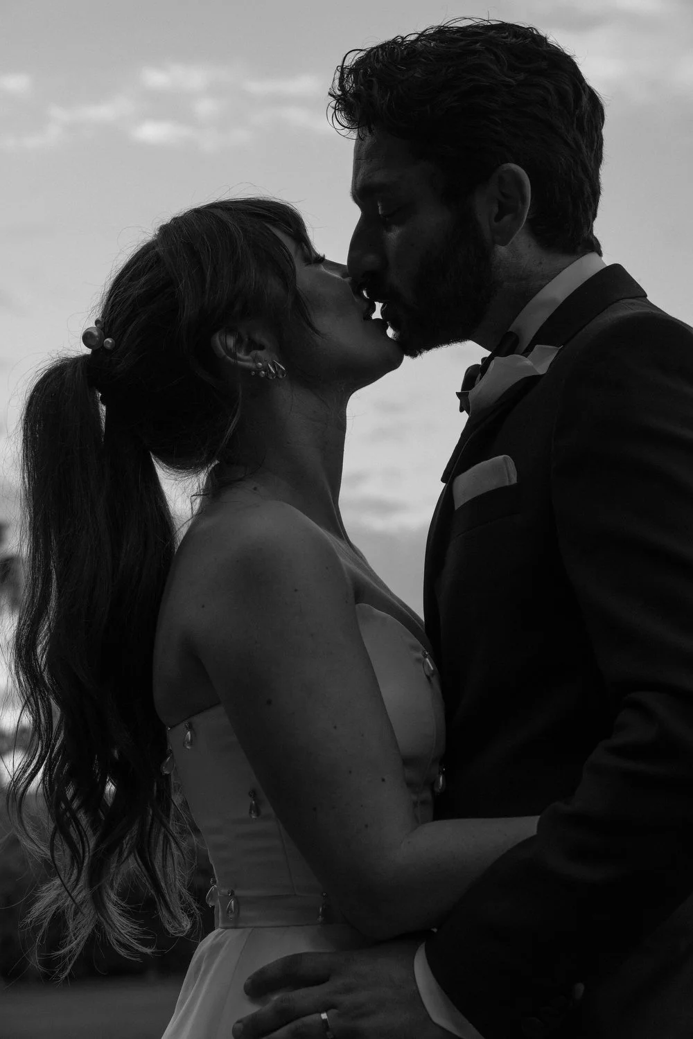 A silhouette of a couple in wedding attire sharing a kiss outdoors, with a cloudy sky in the background.