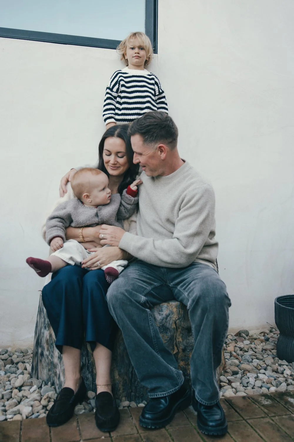 Los Angeles Family photographer. Santa Barbara family photographer. A family of four, including a mother, father, and two young children sitting and standing against a plain wall outdoors.
