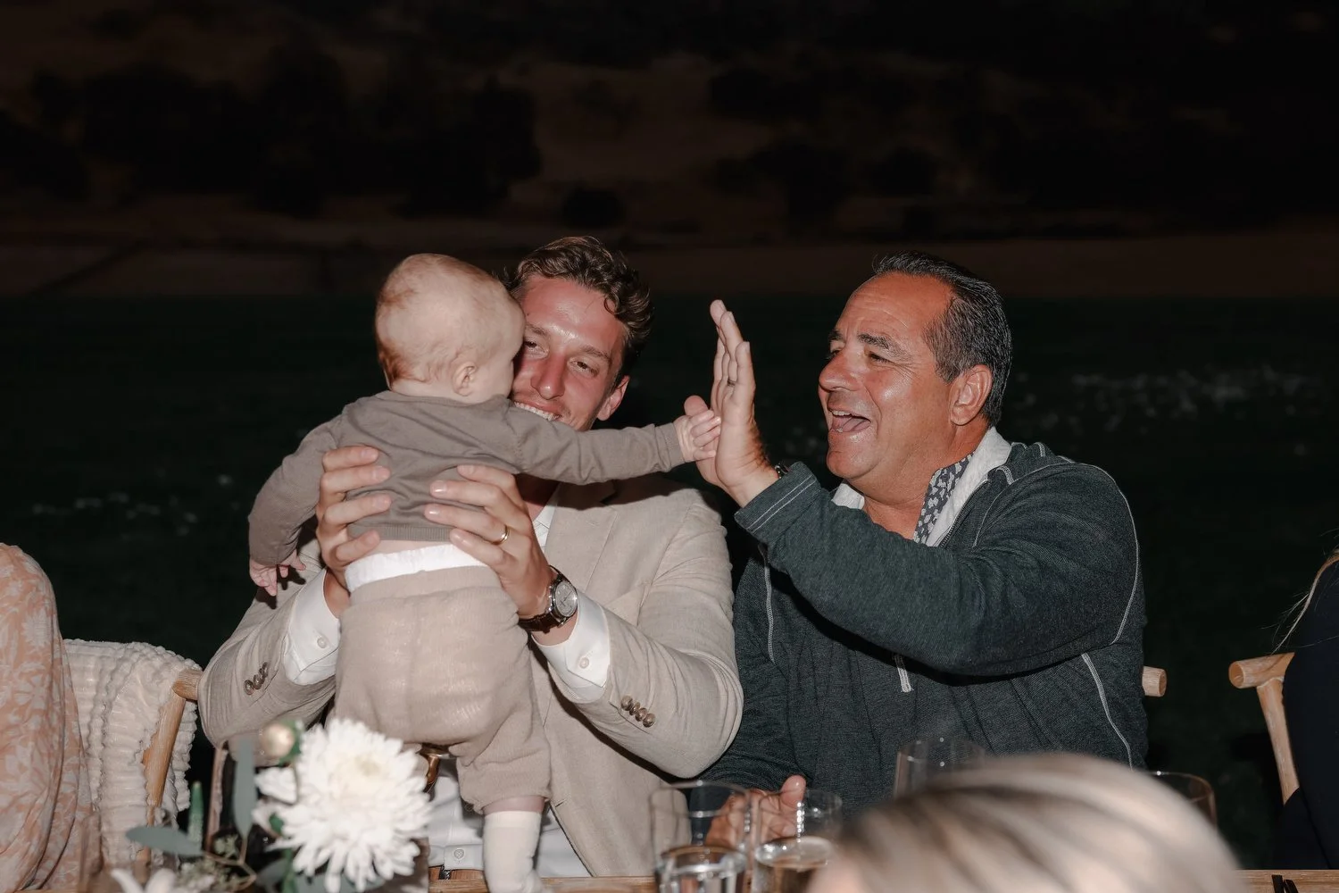 Three generations of men, including a baby, a young man, and an older man, share a joyful moment outdoors at night, with the baby reaching out and giving a high five to the older man.