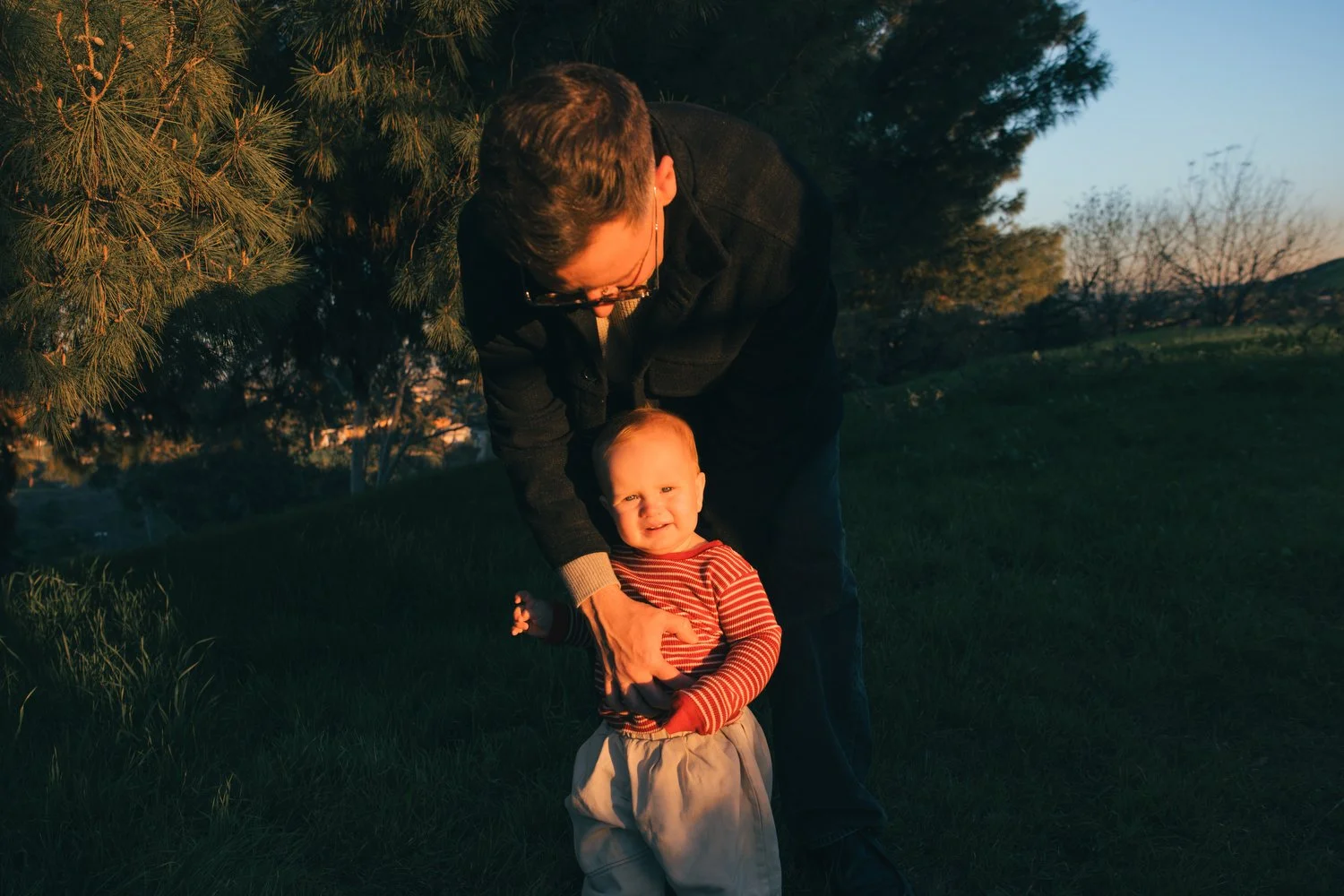 Los Angeles Family photographer. Santa Barbara family photographer.  A man helping a young child stand on a grassy field in the evening with trees in the background.