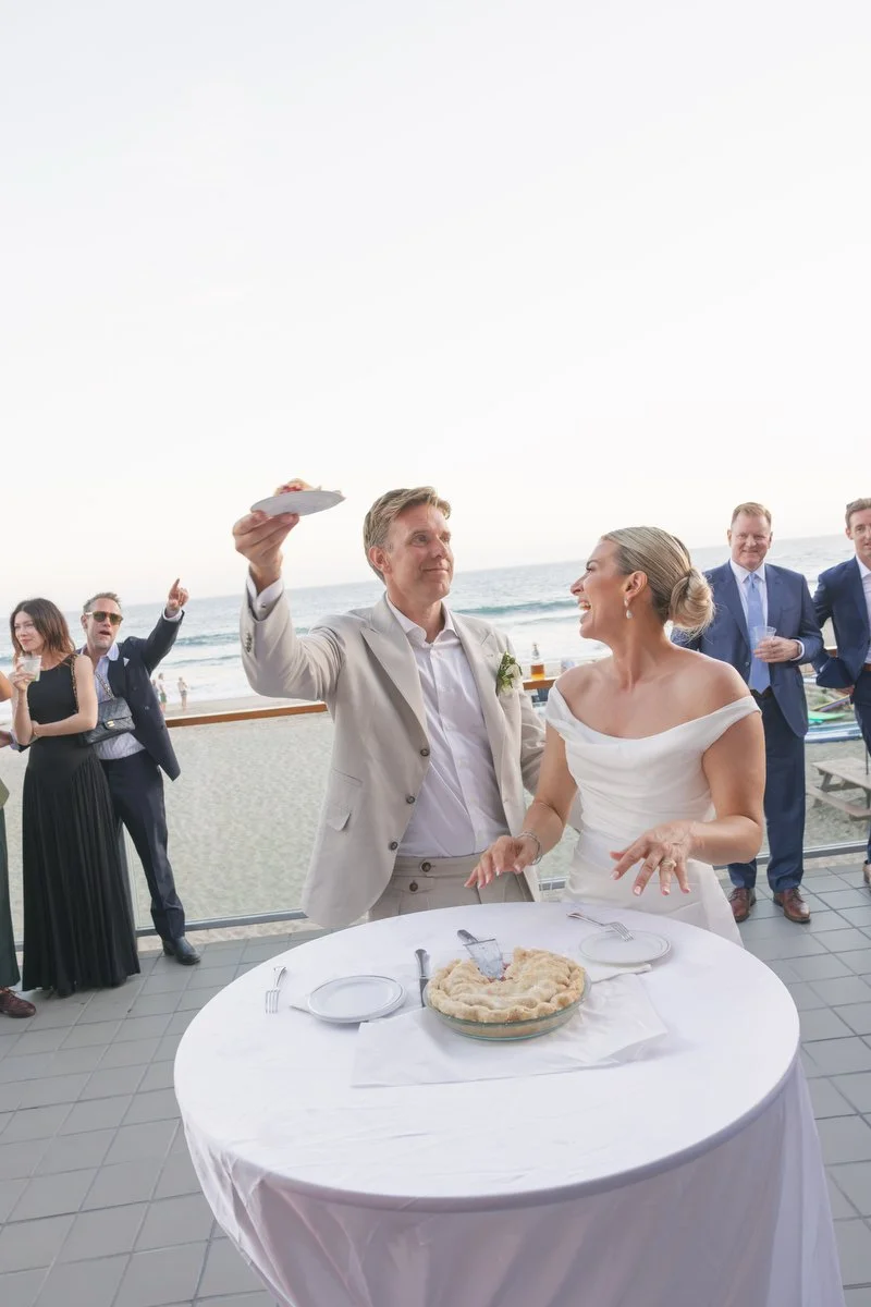 A newlywed couple at a wedding reception by the beach, with the groom holding a plate of cake and footage of guests celebrating in the background. I am a Los Angeles and Santa Barbara wedding photographer. 