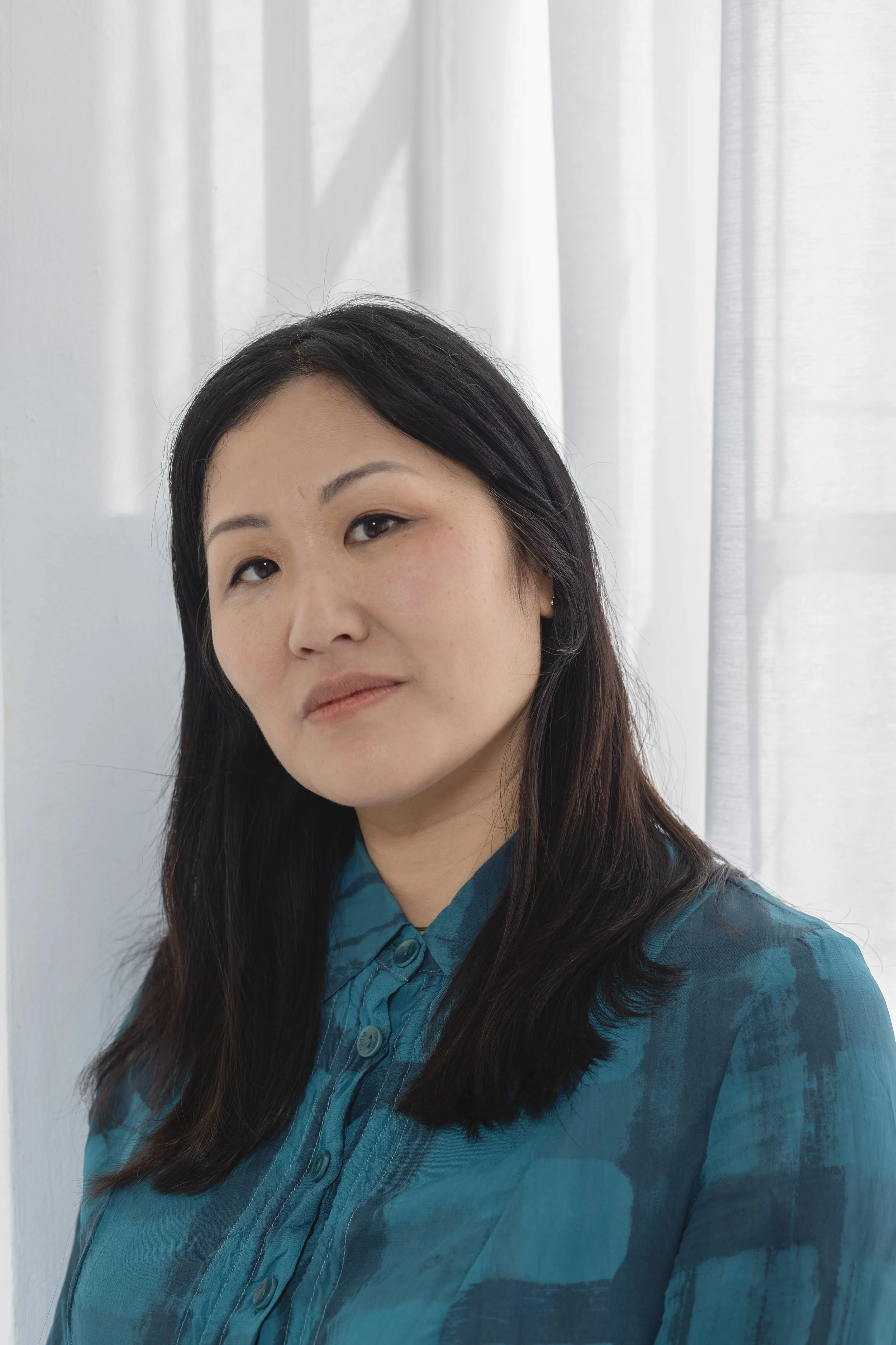 A woman with long dark hair wearing a teal button-up shirt, standing in front of white curtains and a white wall.