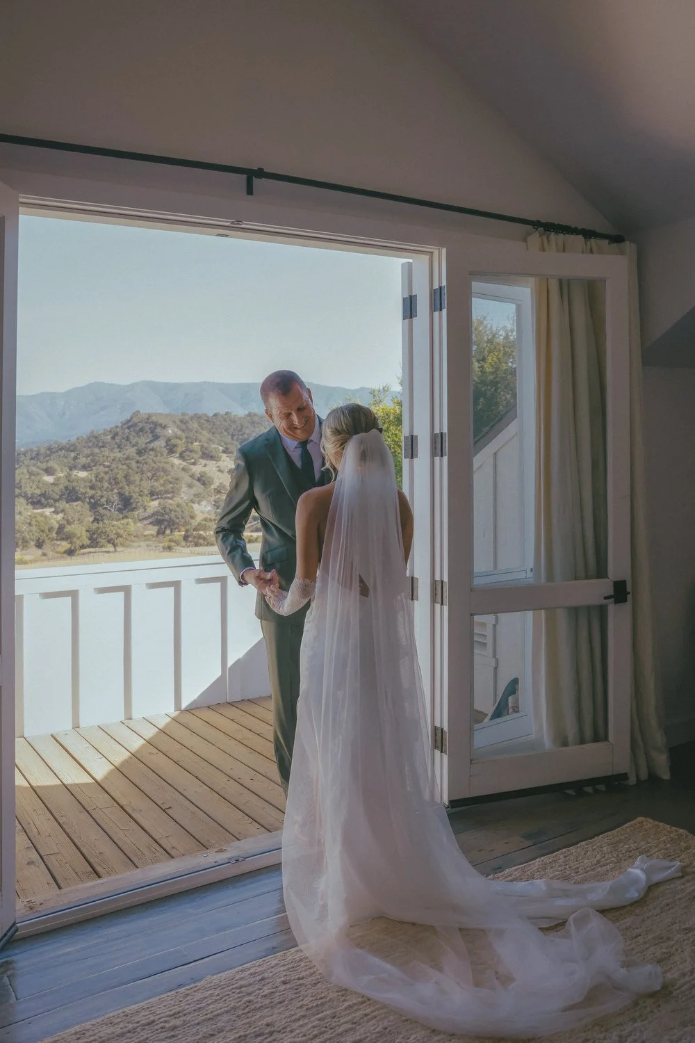 A bride and an older man, possibly her father, stand on a balcony with a scenic mountain view in the background. The bride wears a white wedding dress and veil, and the man is in a suit, holding her hand and smiling.