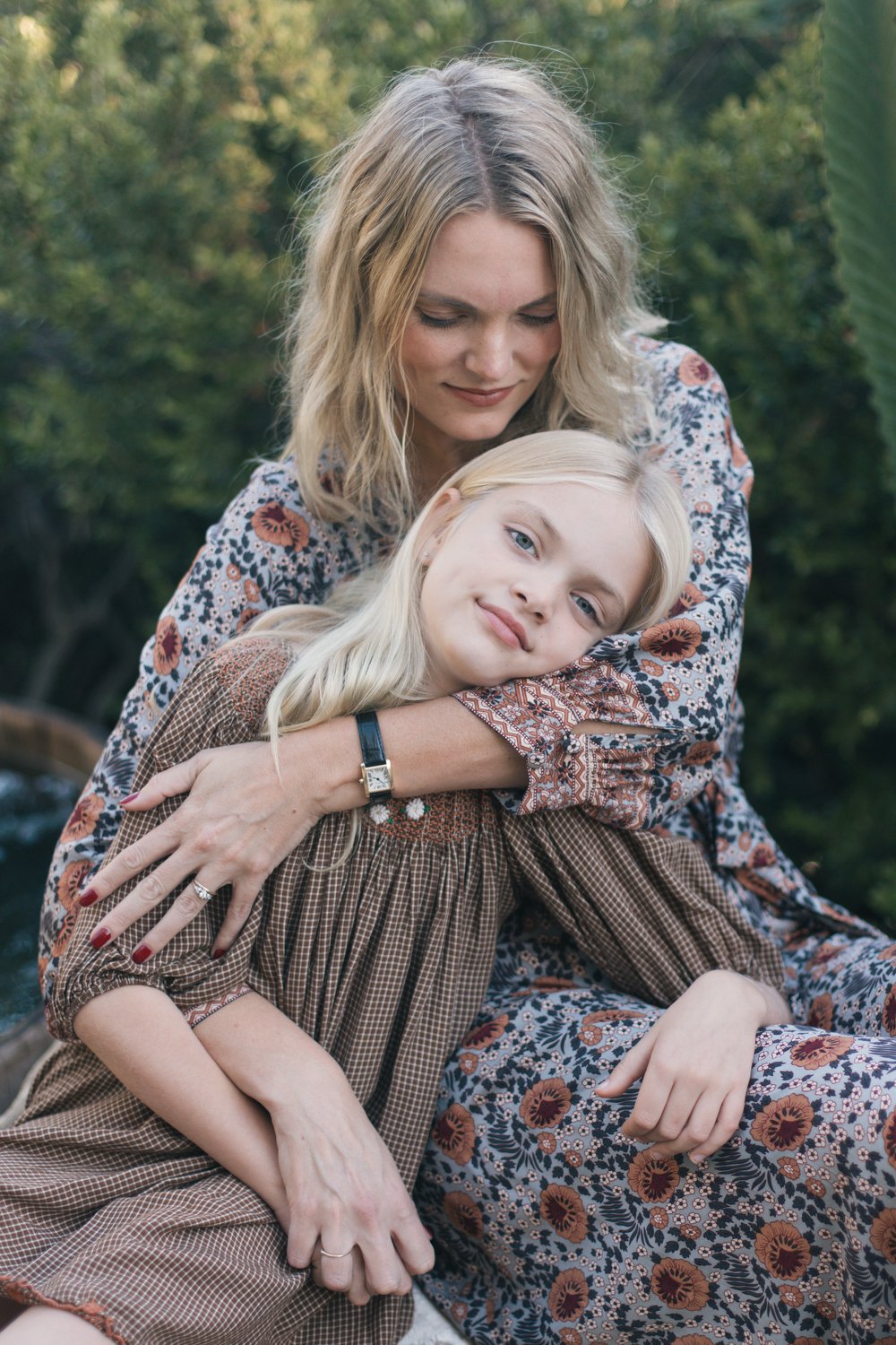 Los Angeles Family photographer. Santa Barbara family photographer. A woman and a young girl sitting outdoors with the woman hugging the girl, both smiling softly, with green trees in the background.