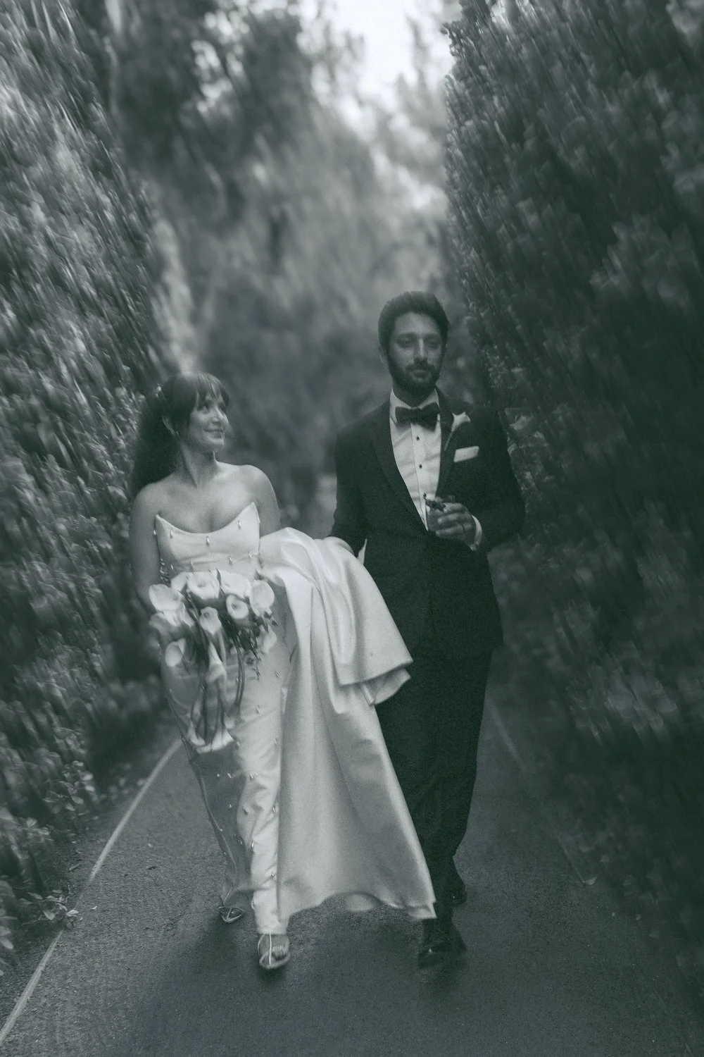 A black-and-white photo of a bride and groom walking together through a narrow outdoor passageway surrounded by tall, dense foliage. The bride is wearing a strapless wedding gown and holding a bouquet, while the groom is dressed in a tuxedo with a bo
