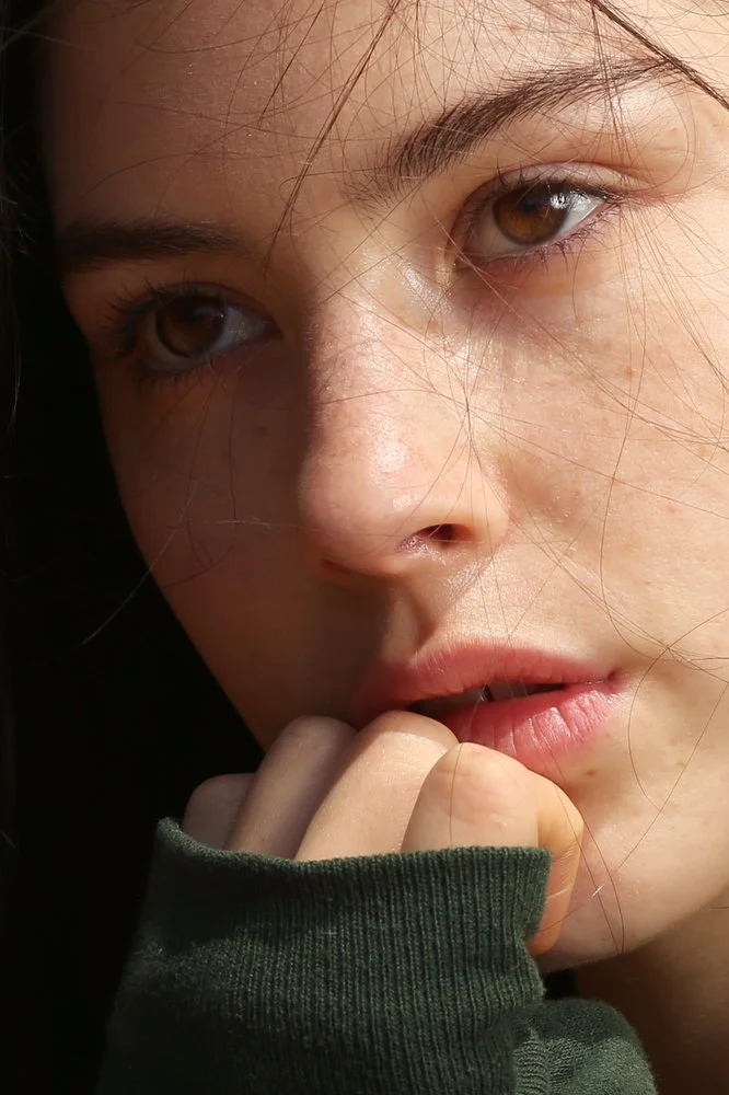 Close-up of a young woman's face with brown eyes, slightly parted lips, and hair falling across her forehead, with her hand near her mouth in a reflective pose. Gracie Abrams photograph. Celebrity photographer. 