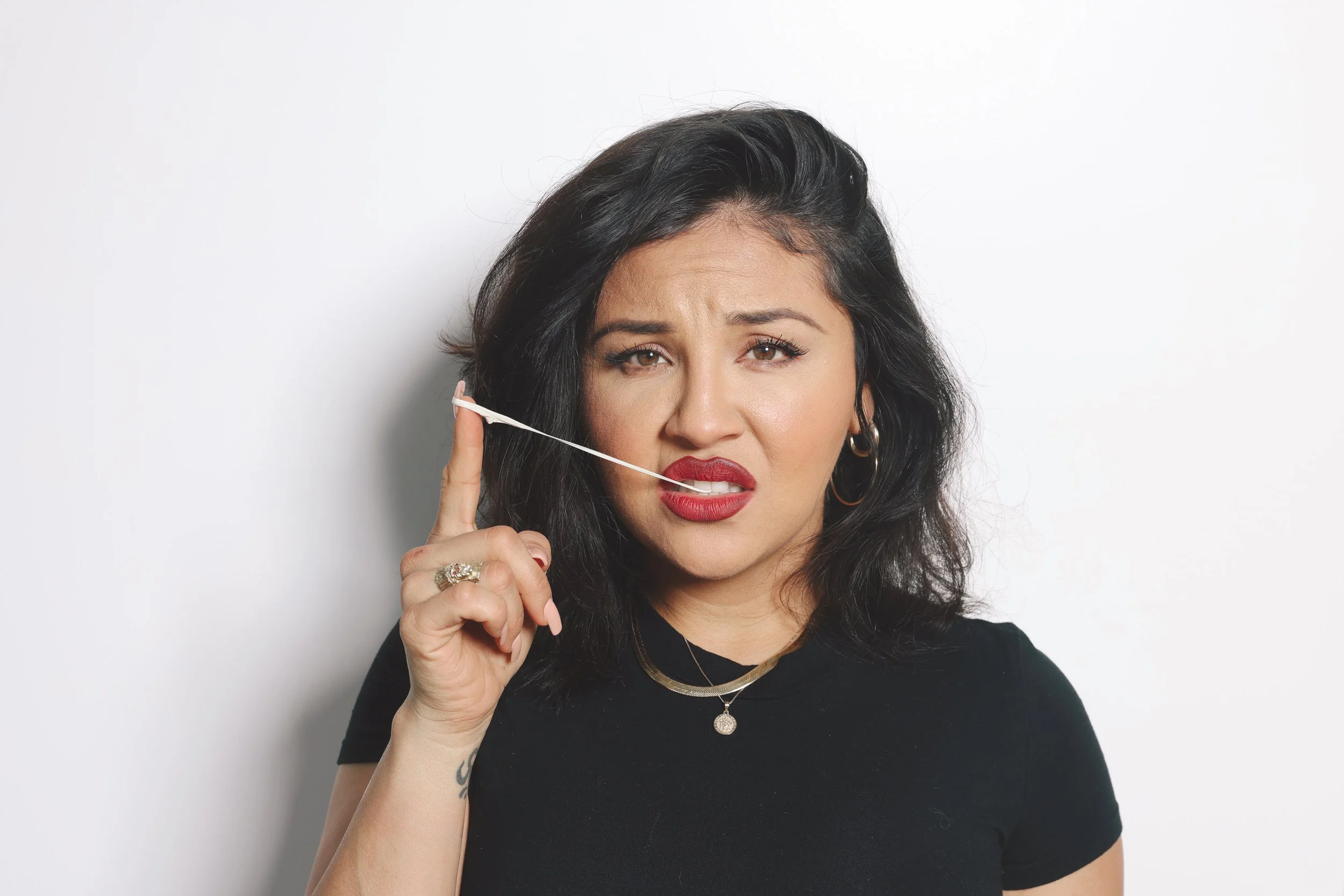A woman with black hair, wearing red lipstick and earrings, holds a white headphone microphone close to her face against a plain white background.