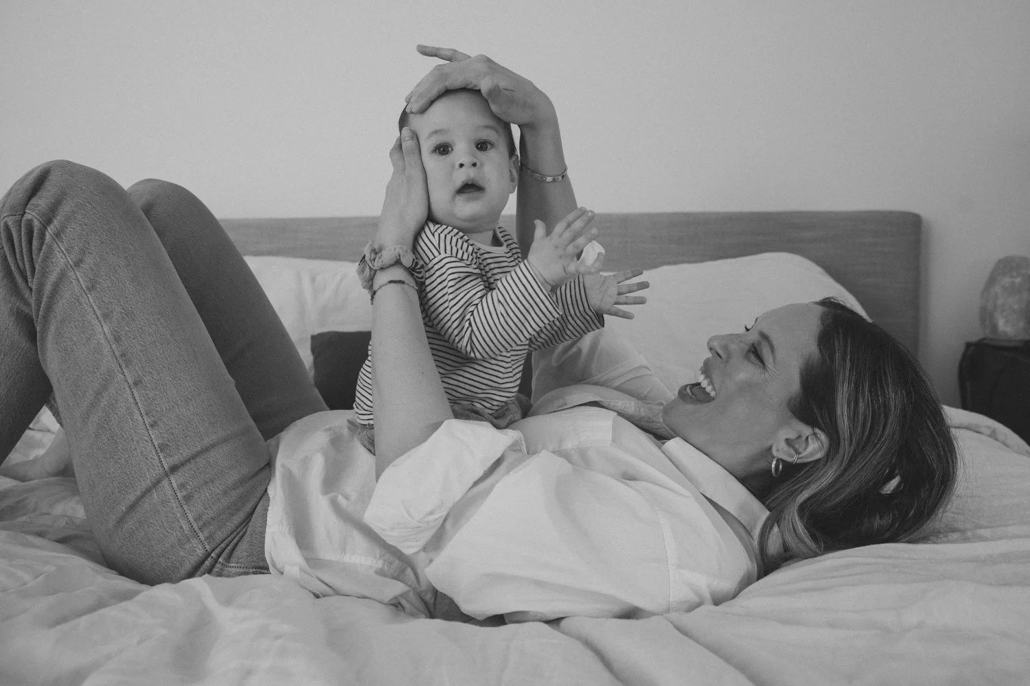 Los Angeles Family photographer. Santa Barbara family photographer.  A woman with long hair and earrings lying on a bed, smiling, holding a young child dressed in a striped shirt, playing and touching his head in a cozy bedroom.