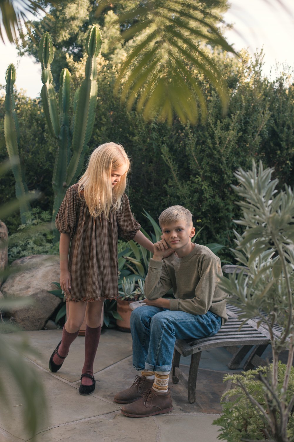 Girl with long blonde hair in a brown dress and a boy with short blonde hair in a beige shirt and blue jeans, seen in a lush garden with various green plants and cacti, with the girl gently touching the boy's shoulder. Los Angeles photographer