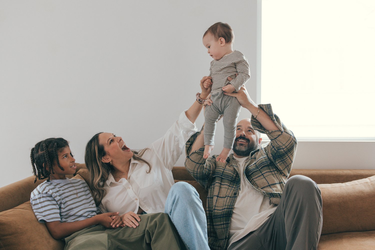 Los Angeles Family photographer. Santa Barbara family photographer. Family of four enjoying time together on a sofa, with a man lifting a toddler, a woman and a girl watching.