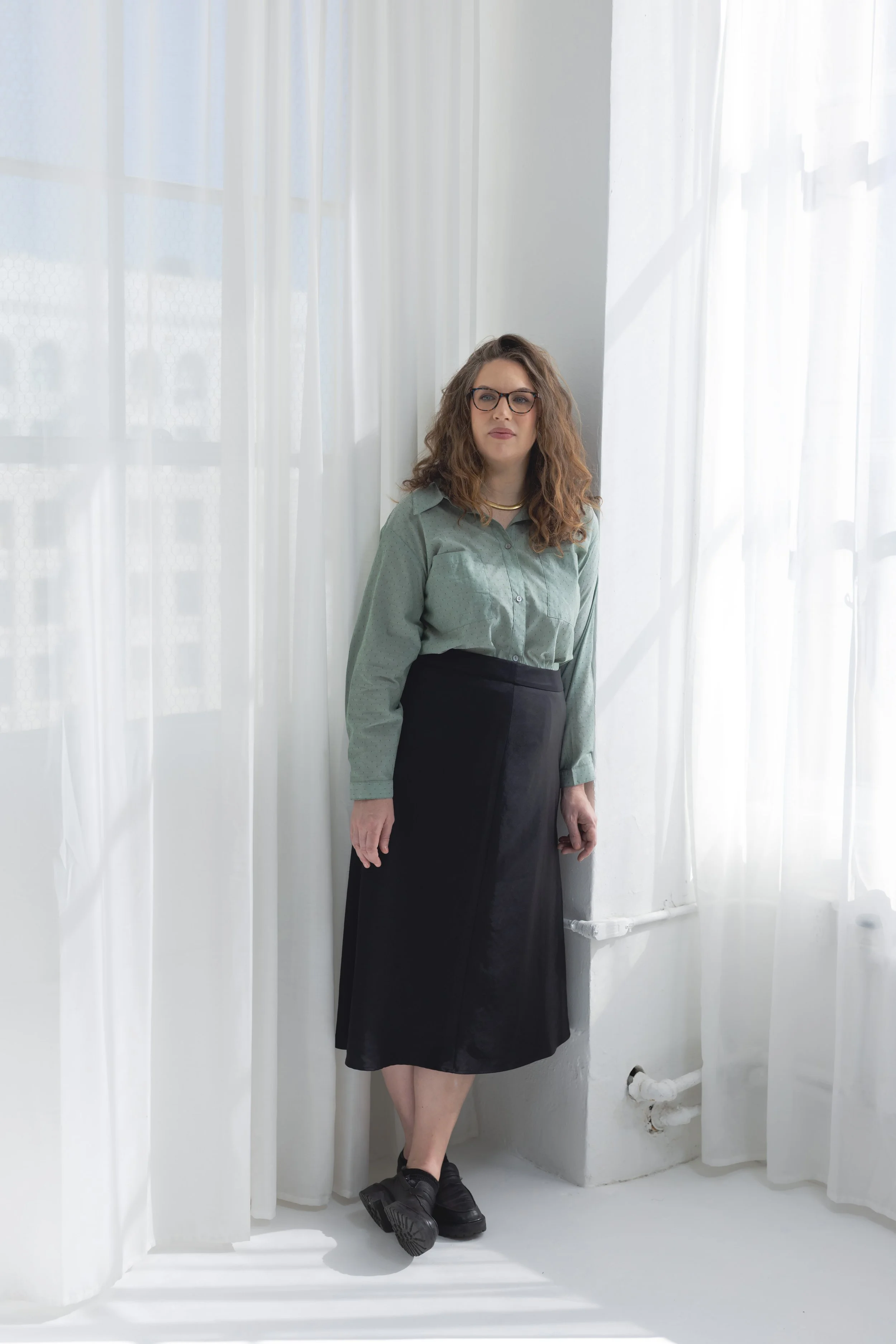 A woman with curly brown hair, glasses, wearing a green button-up shirt, black skirt, and chunky shoes standing against a white curtain backdrop with natural light.