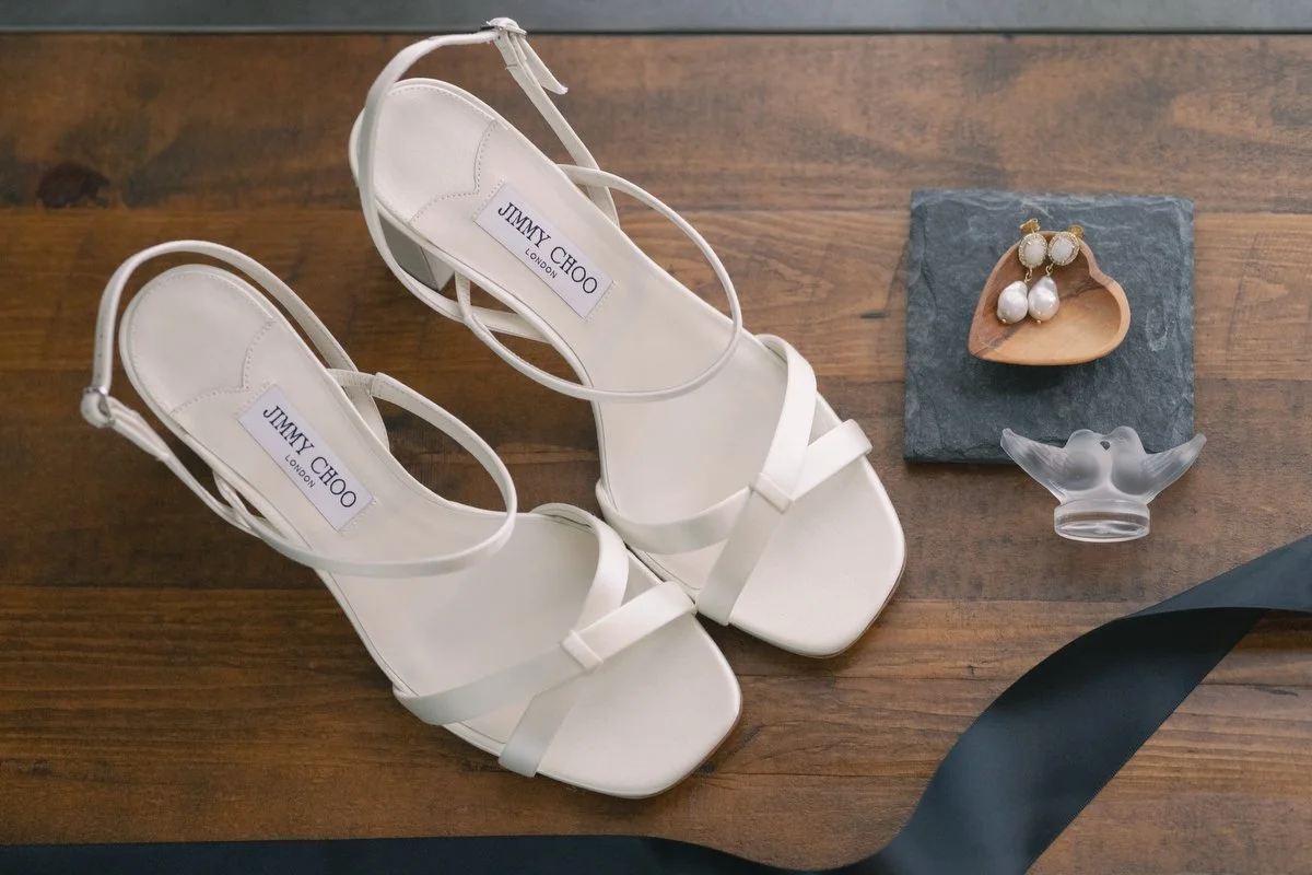 White Jimmy Choo high-heeled sandals with ankle straps, a wooden dish with pearl earrings, a grey glass bird figurine, and a black ribbon on a wooden surface.  I am a Los Angeles and Santa Barbara wedding photographer. 
