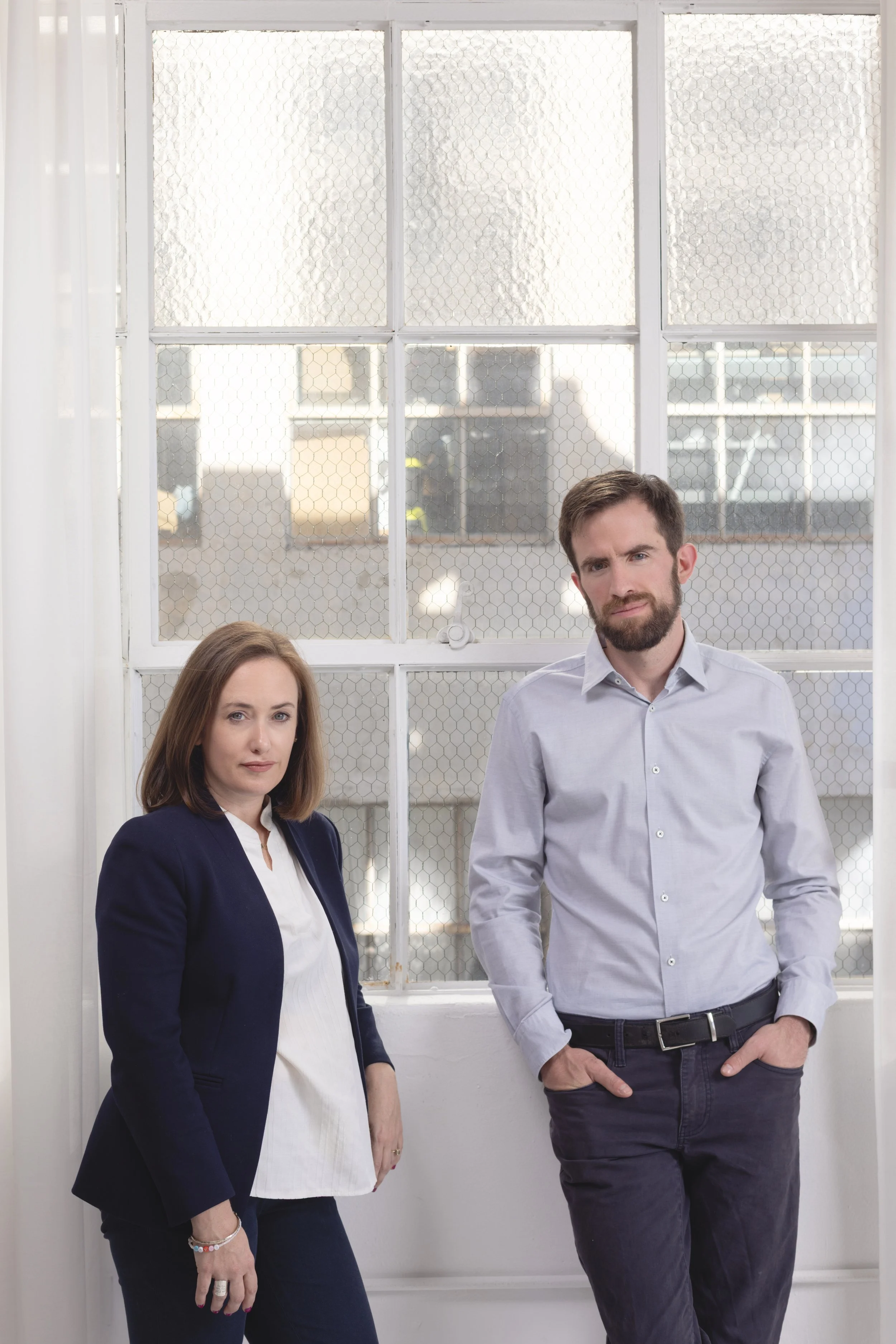 A woman with straight, shoulder-length brown hair wearing a navy blazer and white blouse, and a man with short brown hair and beard wearing a light gray dress shirt, standing in front of a window with wire mesh, with buildings visible outside.