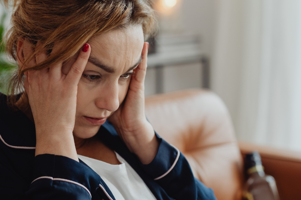 A woman with reddish hair, holding her head with both hands, appears distressed or worried, sitting on a beige couch in a home setting.