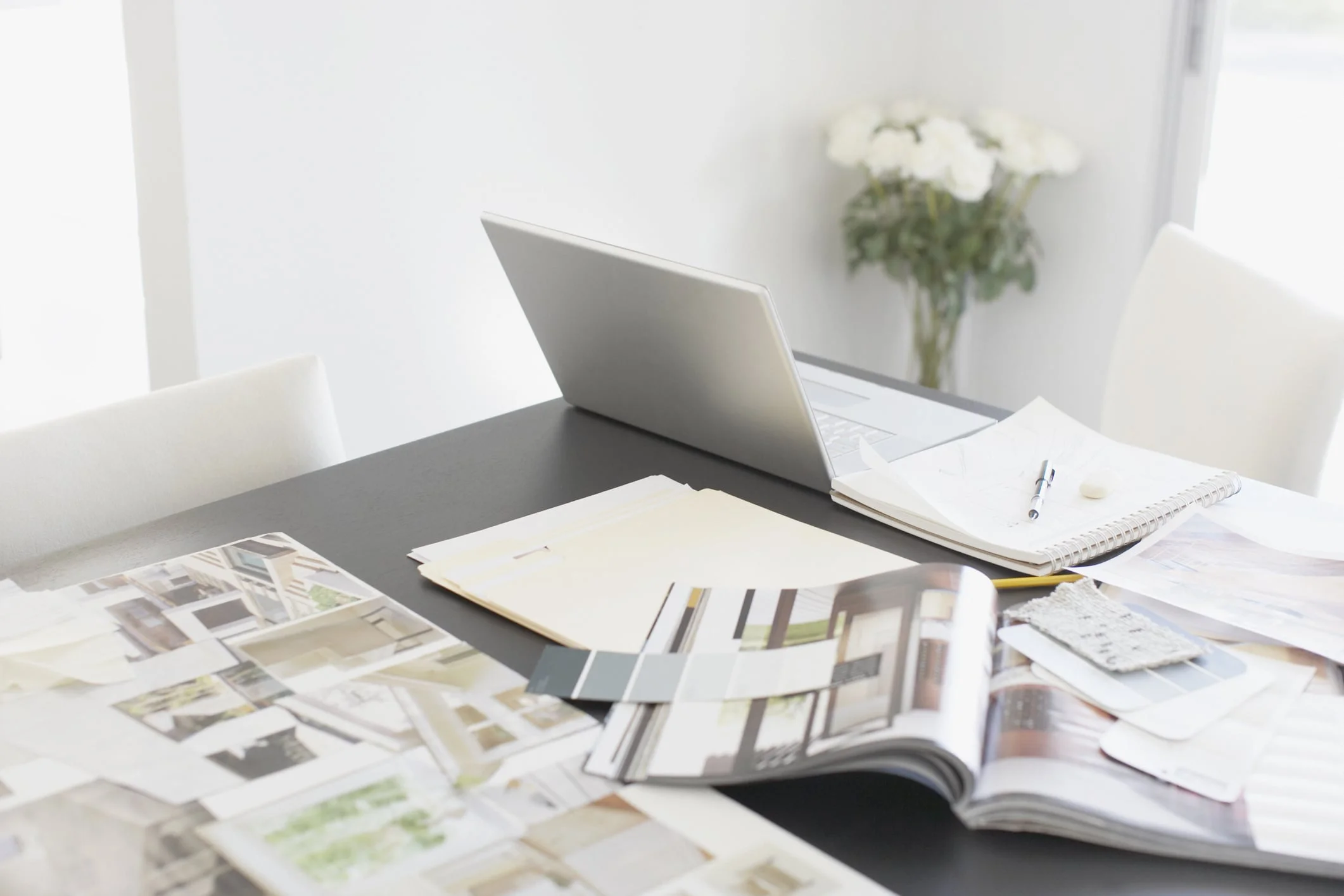 Dark, modern workspace table in white room with laptop, folders, swatches, lookbooks and notebook. White roses sit in corner.
