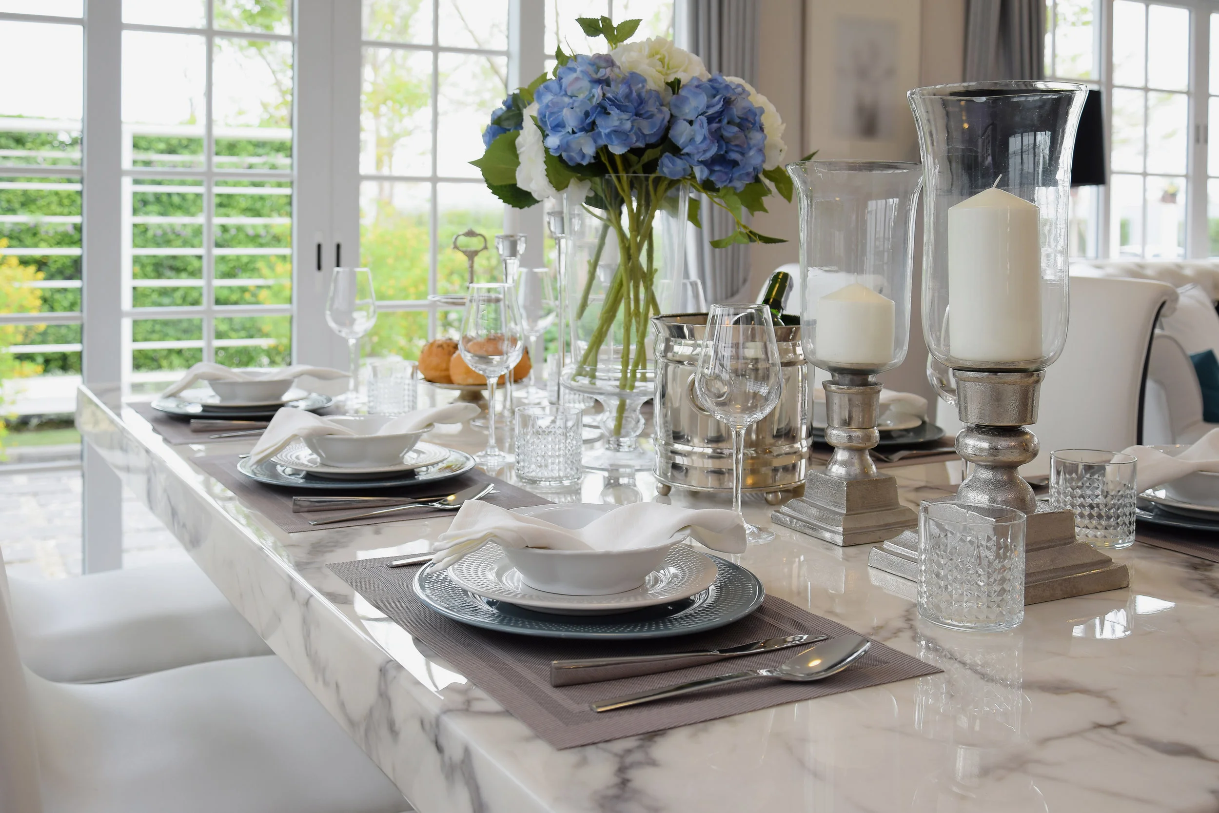 Gleaming white marble table set for a meal with hydrangea and candlestick accents, illuminated by light from nearby windows.