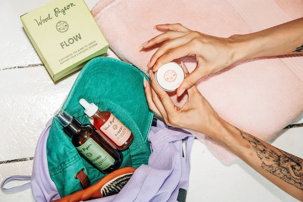 A person's hands holding a small container of Wood Pigeon product, surrounded by skincare items including a spray bottle, a dropper bottle, a wooden brush, and a box of Wood Pigeon FLOW tea, with a pink towel underneath. A backpack with cloth material and a lavender fabric are also visible.