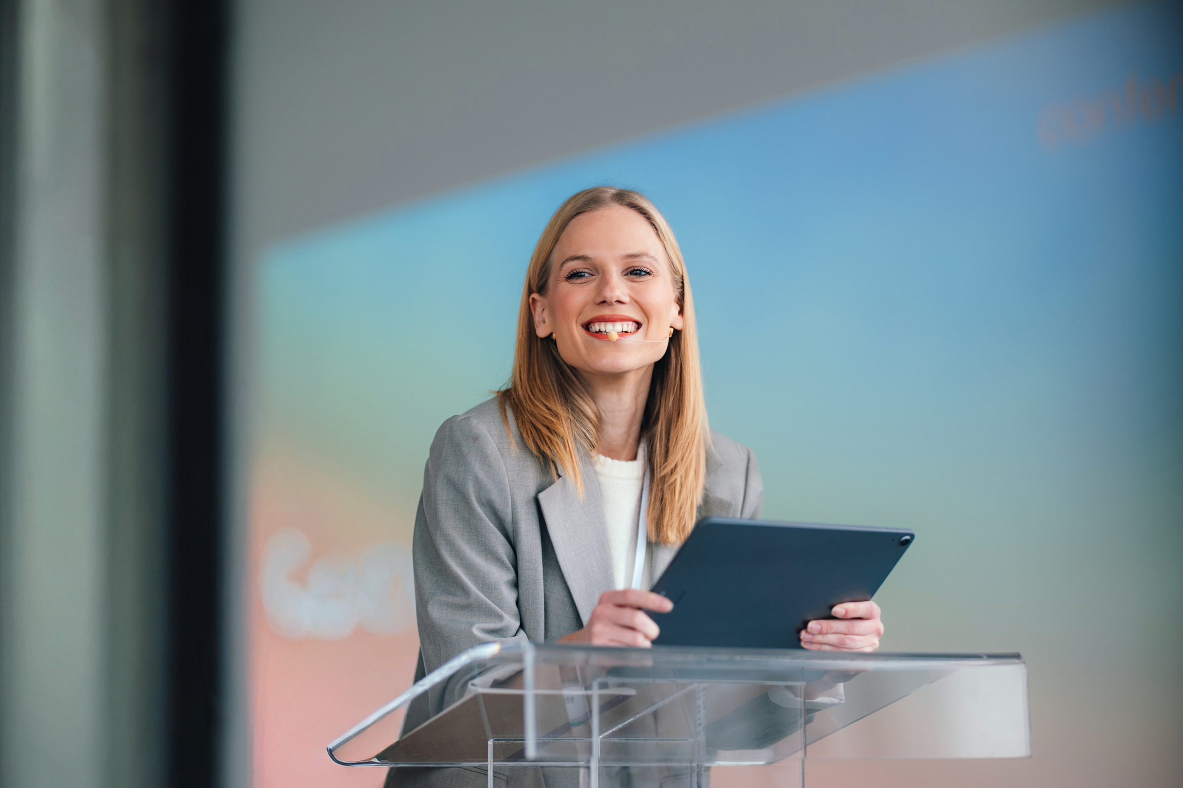 A woman in a gray blazer smiling and holding a tablet while standing at a podium, with colorful background behind her.