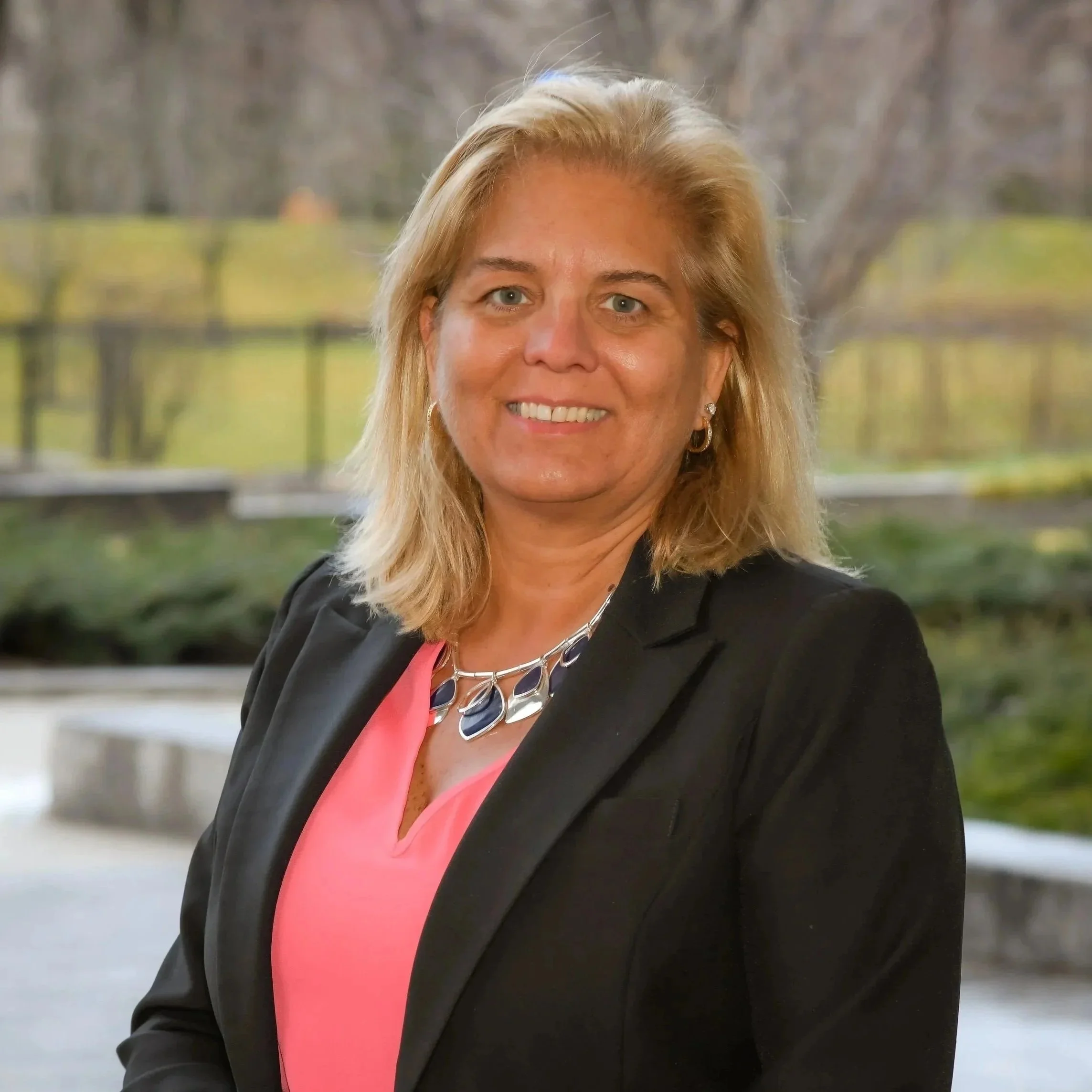 A smiling woman with shoulder-length blond hair, wearing a black blazer, pink blouse, and silver jewelry, standing outdoors with blurred trees and a pond in the background.