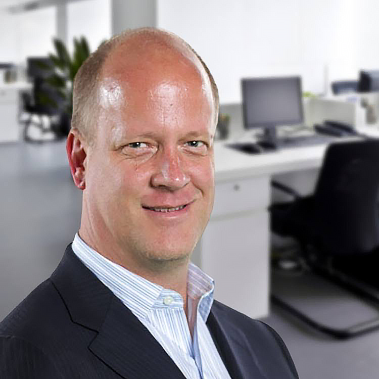 A man in a business suit smiling in an office setting with desks, monitors, and chairs in the background.