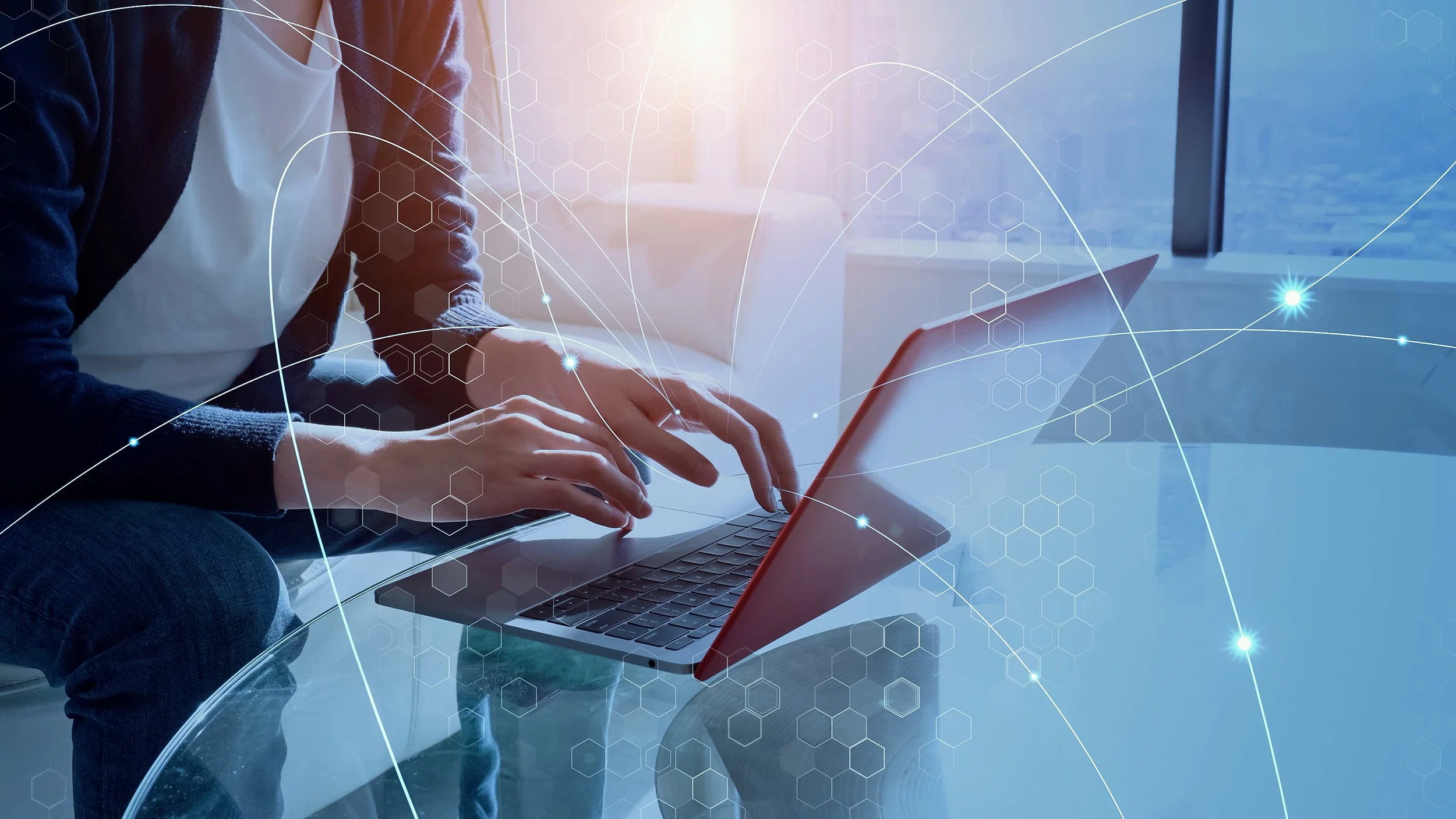 A person using a laptop on a glass table with digital graphics overlay, including hexagons and lines, suggesting technology or digital connectivity, in a modern, well-lit space with large windows.