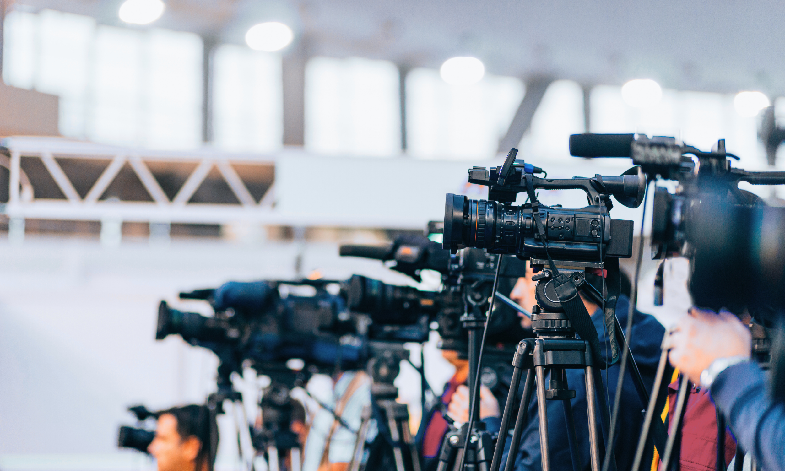 Multiple professional video cameras set up on tripods at a press conference or media event in a well-lit indoor space.