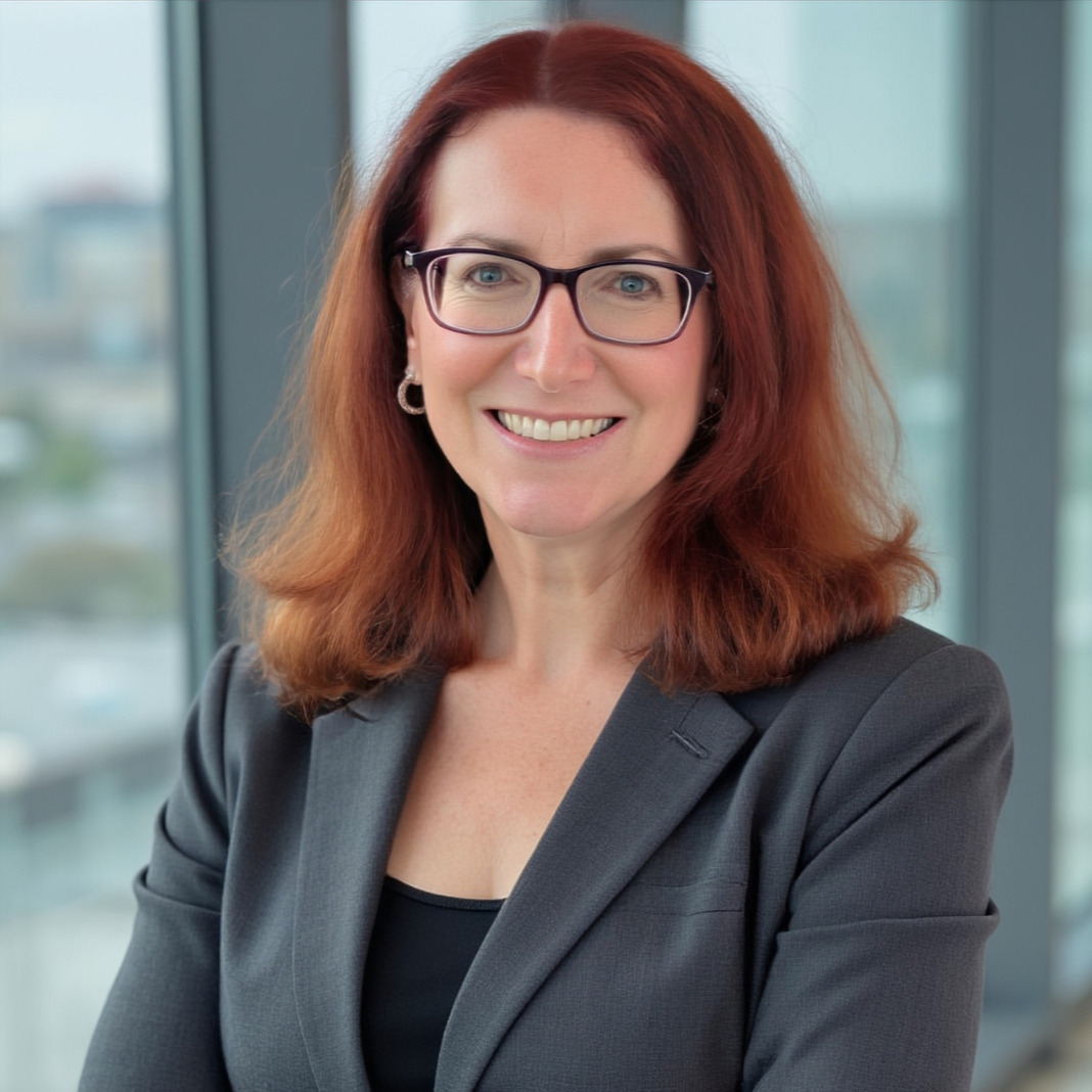 Professional woman with red hair, glasses, and earrings, smiling in an office with large windows in the background.
