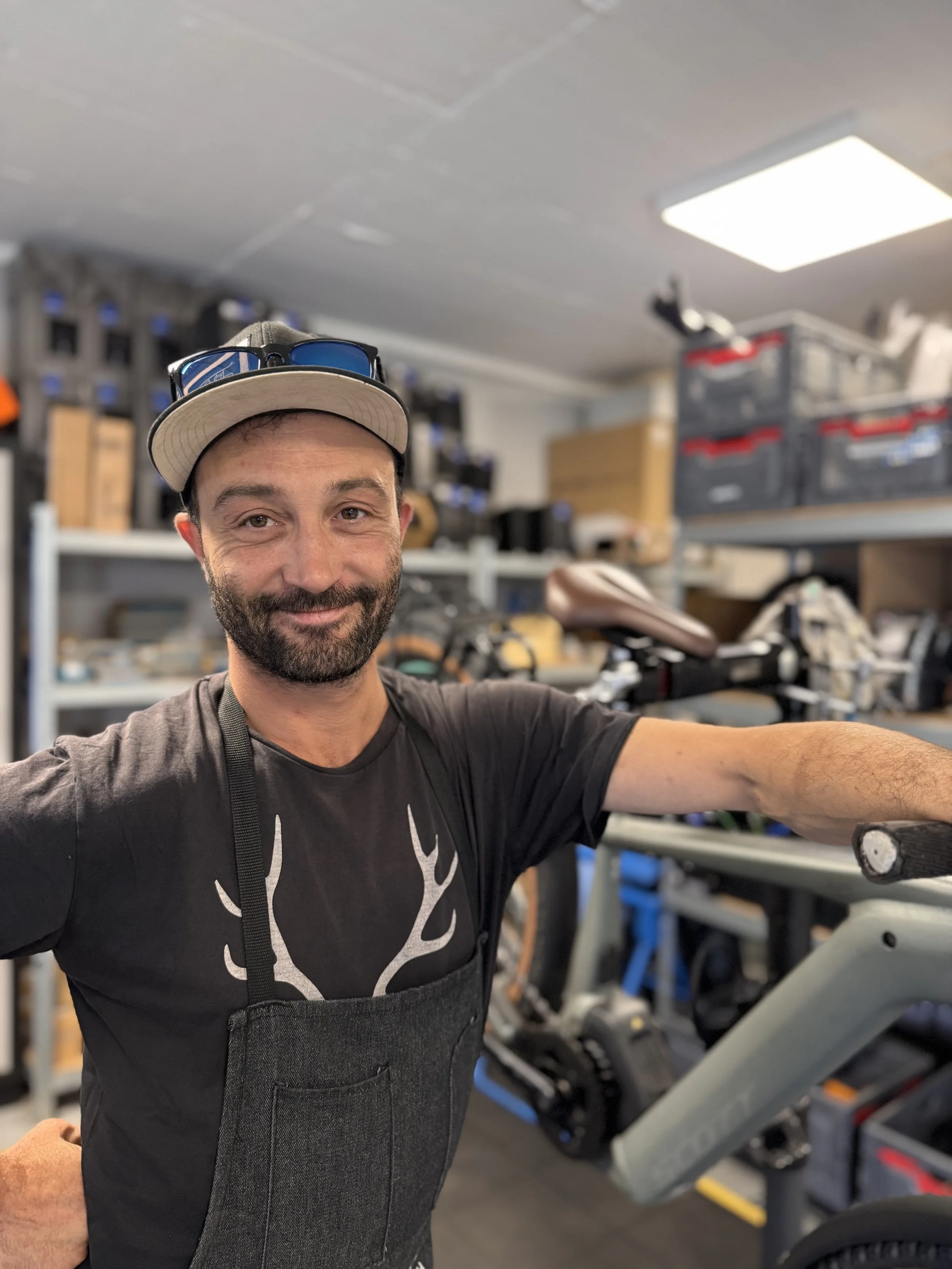 A man with dark hair, beard, and a smile, wearing a beige cap with sunglasses on top, a black t-shirt with a white reindeer silhouette, and a black apron, is taking a selfie in a bike shop. The background shows shelves with bike parts and boxes.