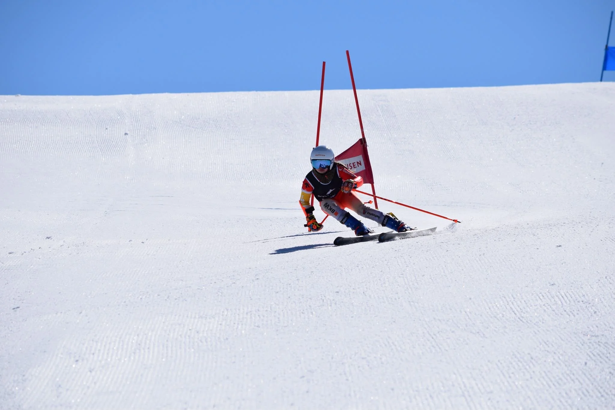 A skier wearing a helmet, goggles, and a racing suit is navigating around a slalom pole on a snow-covered slope under a clear blue sky.