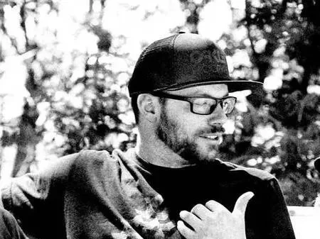 Three people sitting closely together outdoors, engaging in conversation. The man on the left has glasses, a beard, a cap, and is wearing a dark shirt. The woman in the middle is wearing a dark cap, has curly hair, and is wearing a T-shirt. The young man on the right has short hair, tattoos on his arm, and is wearing a light-colored shirt.