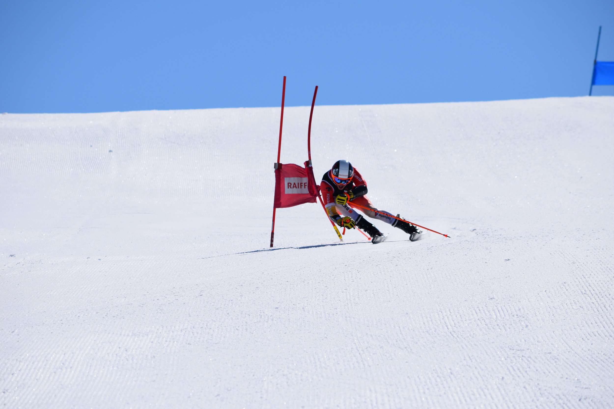 A skier racing down a snowy slope, navigating around a red slalom gate with a blue flag in the background under a clear blue sky.