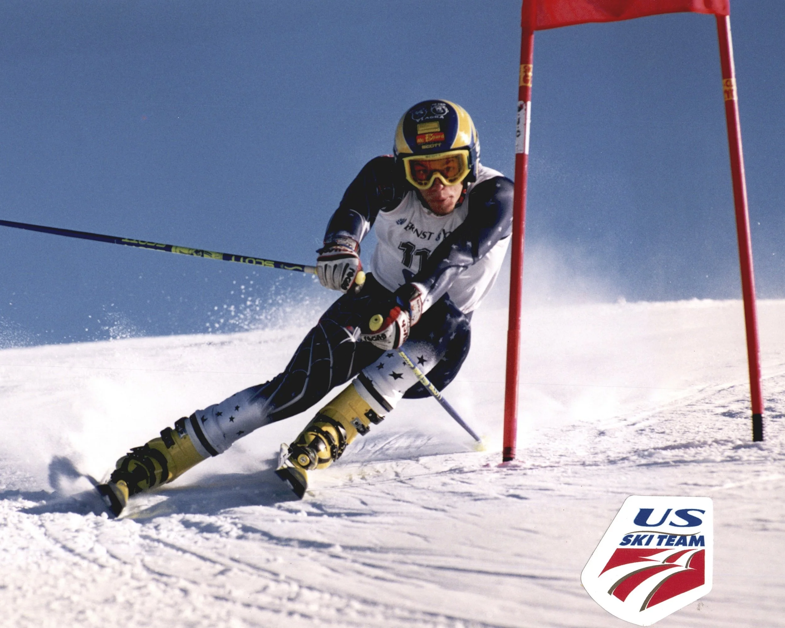 A skier navigating a slalom course on snow, wearing a helmet and goggles, with a red slalom gate in the background and a US Ski Team sign on the snow.